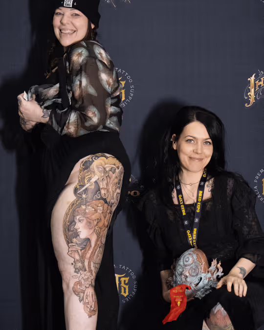 Two women posing at a tattoo event, one showing a large colorful tattoo on her thigh, the other holding a detailed sculpted art piece with a red ribbon.