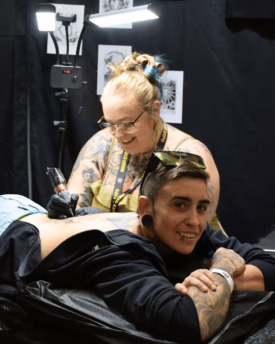 Tattoo artist with glasses and tattoos working on a smiling client's back in a studio with bright lights.