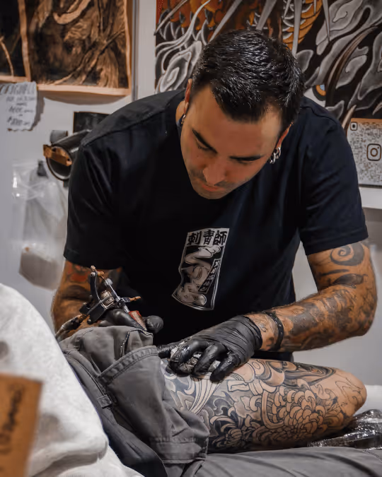 Tattoo artist with gloves working on a detailed black and gray tattoo on a person's leg in a studio.