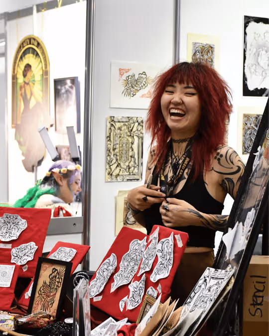 Smiling woman with red hair and tattoos standing behind a table displaying tattoo stencil designs and artwork.