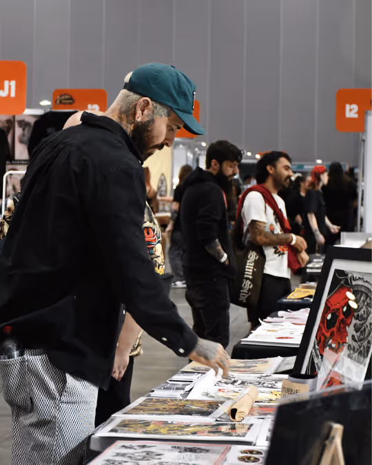 Man with tattoos and a green cap browsing art prints displayed on a table at an indoor market or convention.