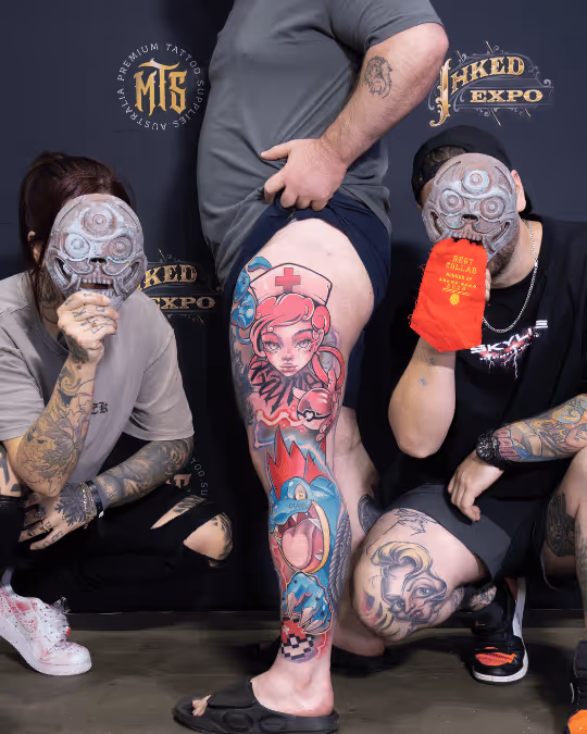 Three tattooed individuals pose in front of a black backdrop with 'INKED EXPO' text; the person in the center shows a vibrant full-leg tattoo featuring a nurse and cartoon character.