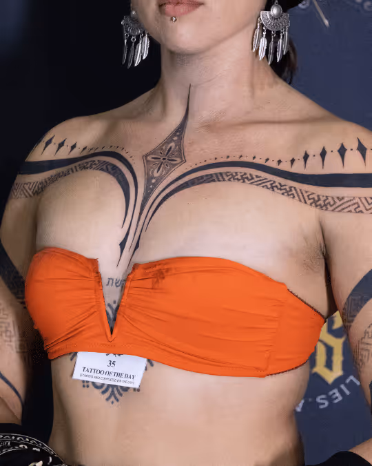 Woman wearing an orange strapless top showing black symmetrical geometric and tribal tattoos across her chest, shoulders, and upper arms, along with large silver earrings and a labret piercing.