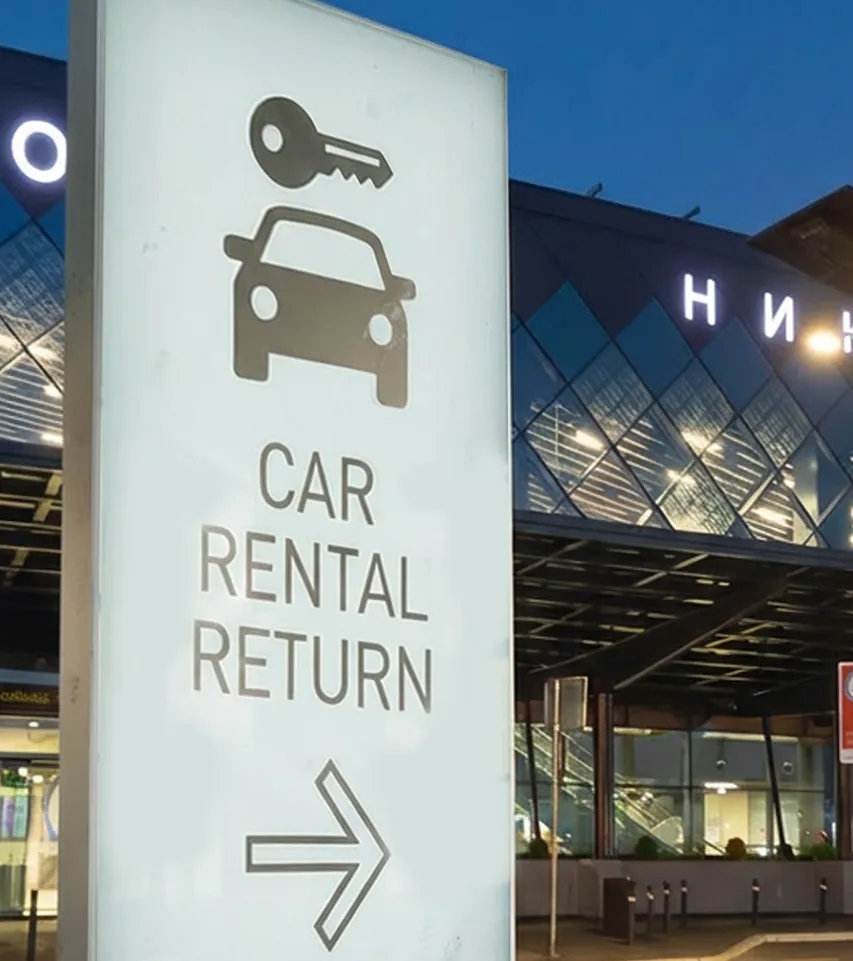 Illuminated sign with a car and key icon indicating car rental return to the right, outside a modern building at dusk.