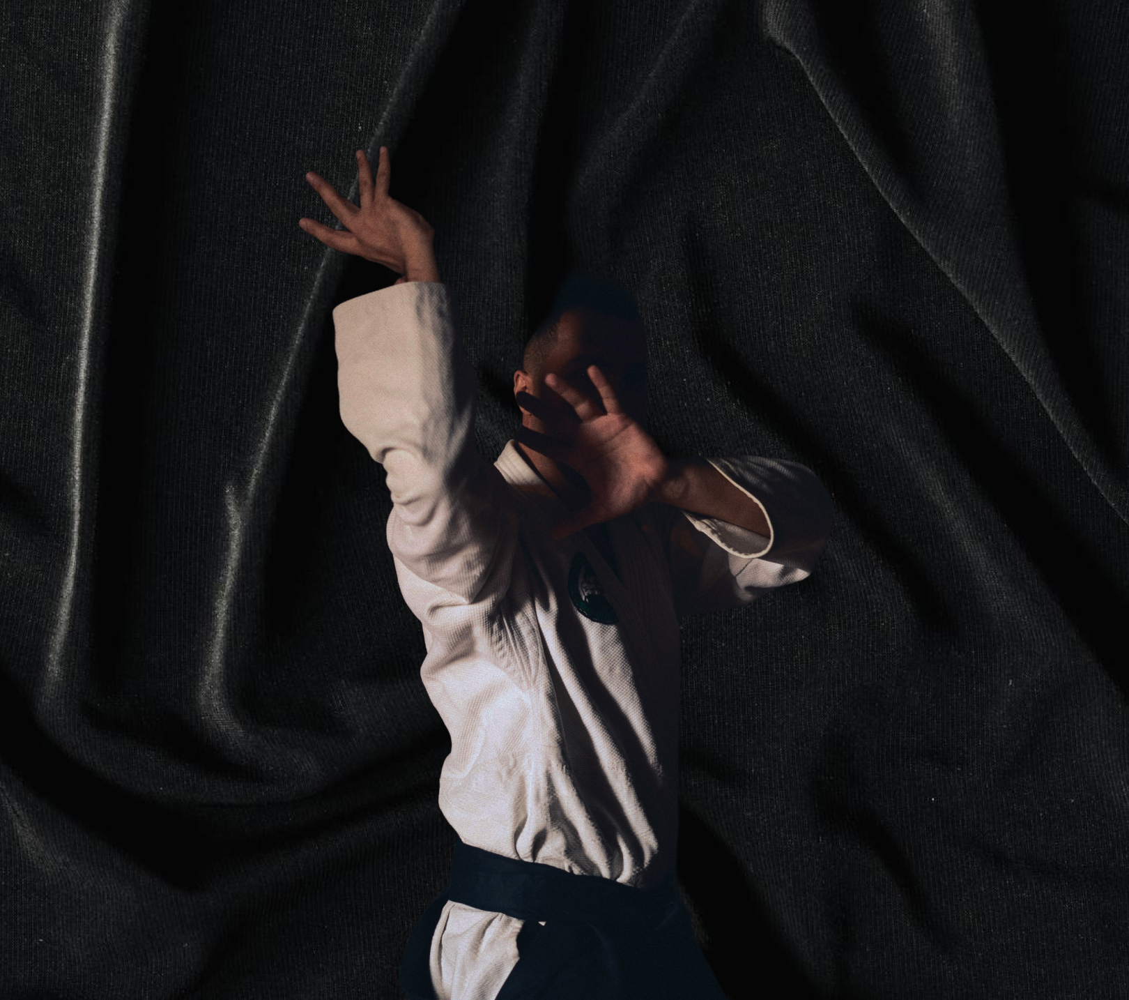 Person in martial arts uniform with a black belt striking a defensive pose against a dark textured fabric background.