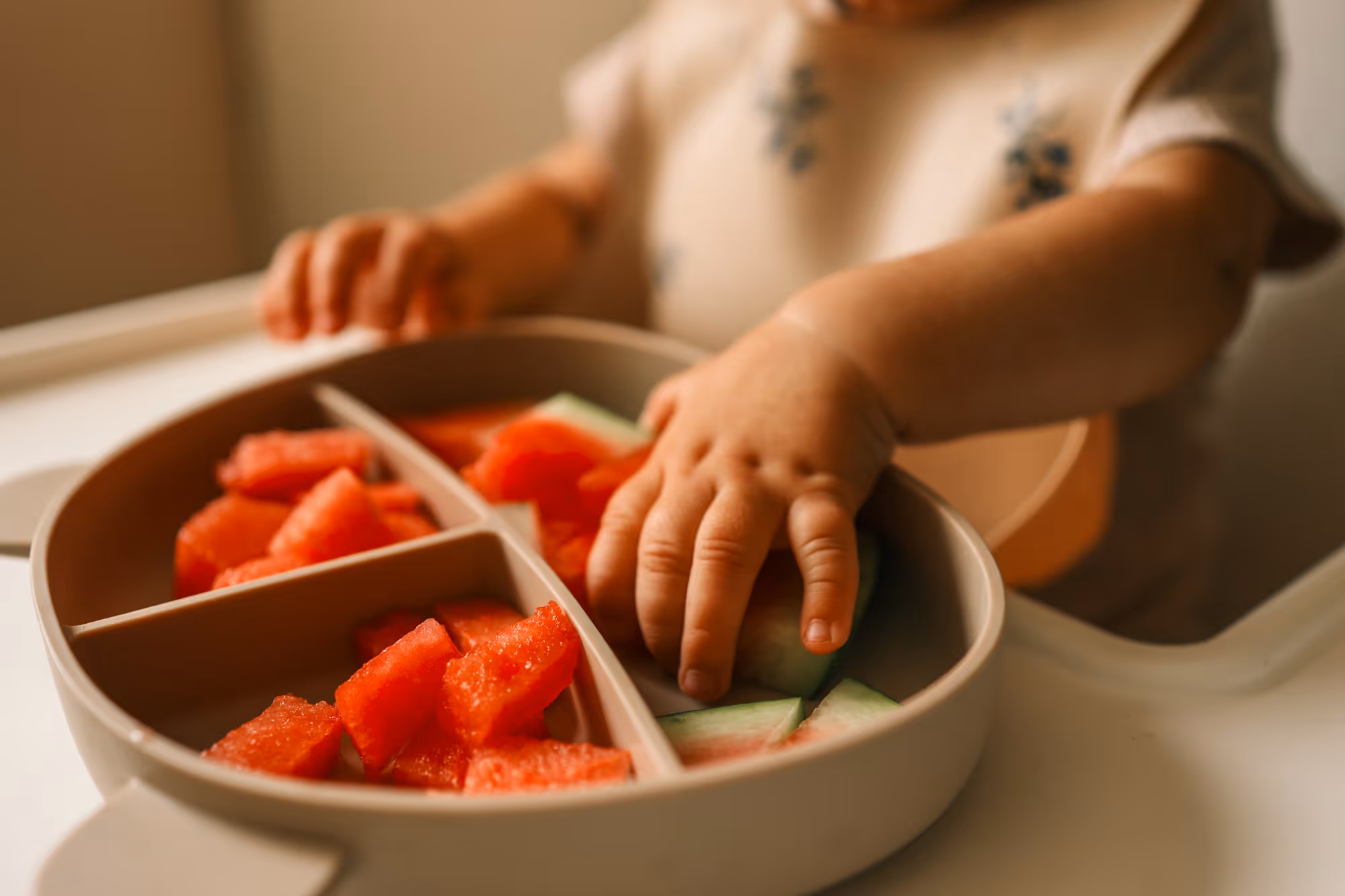 A child’s hand reaching for sliced watermelon pieces in a divided plate on a high chair tray.