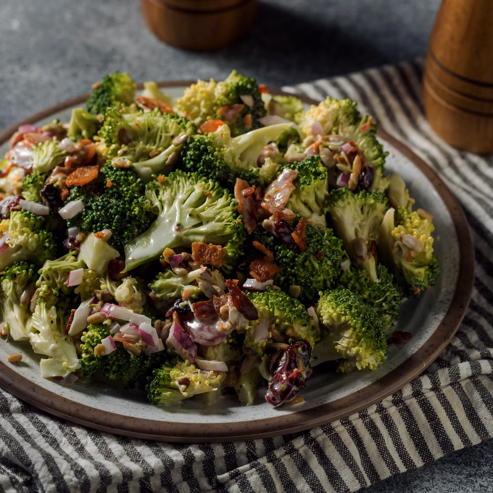 Plate of broccoli salad with bacon bits, red onion, sunflower seeds, and dried cranberries on a striped cloth.