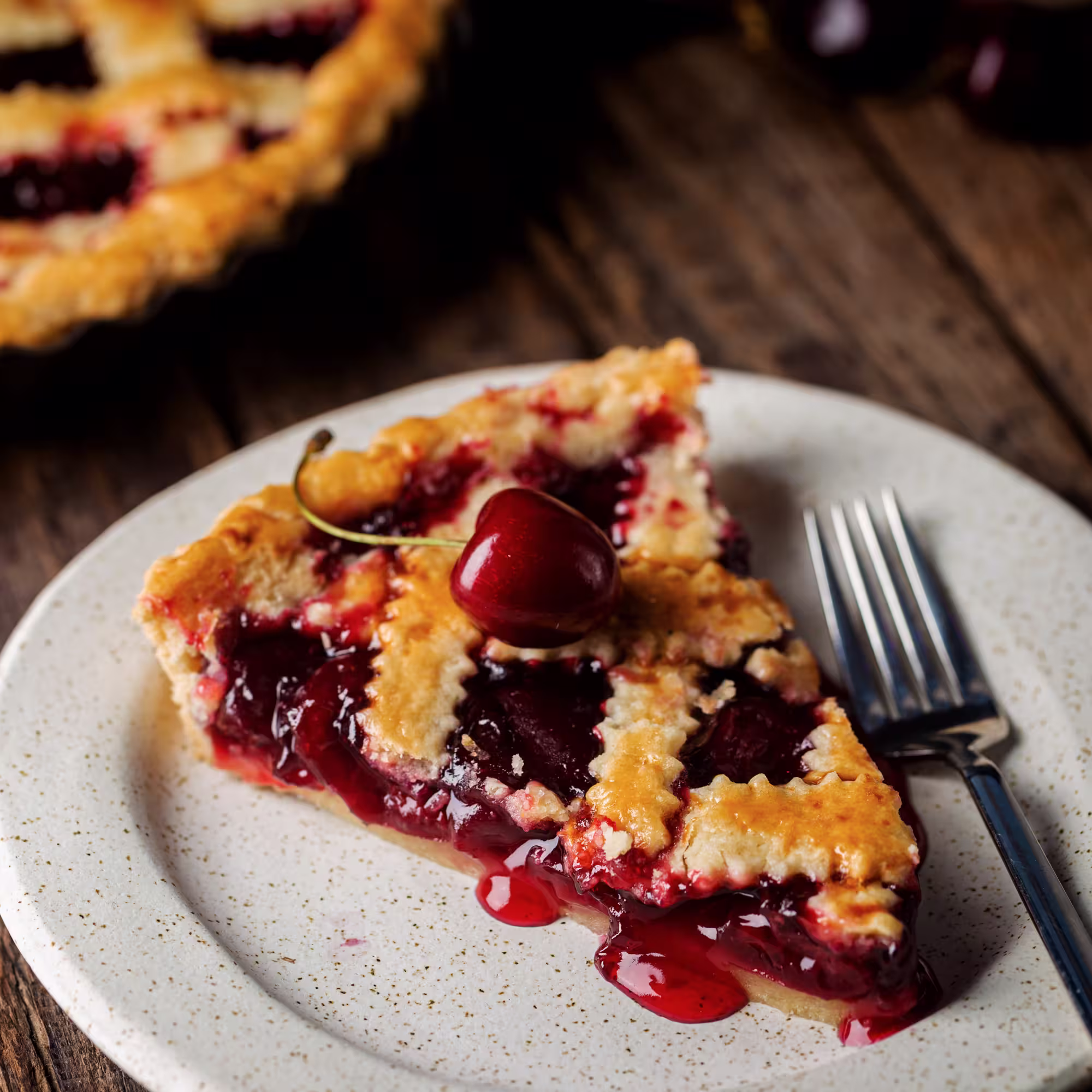 Slice of cherry pie with lattice crust and a cherry on top on a white plate with a fork.