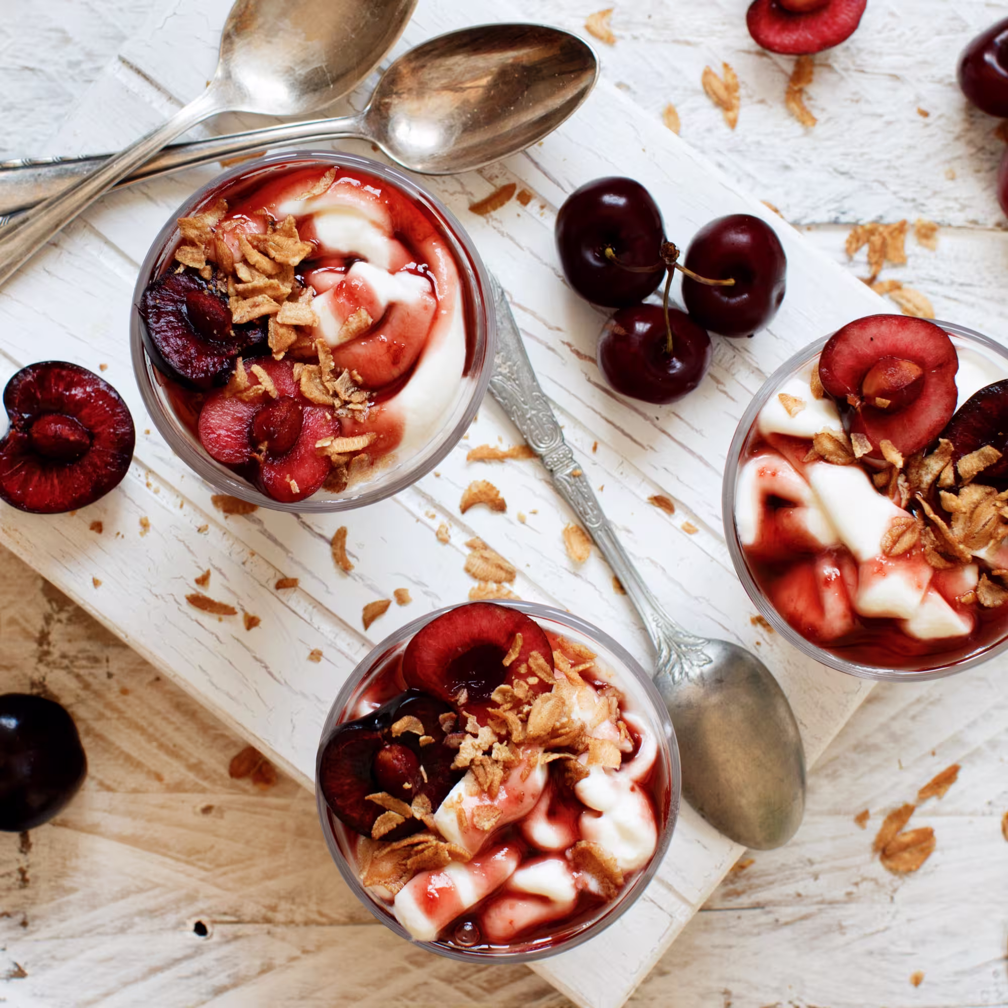 Three glass bowls of yogurt topped with cherry halves and granola on a white wooden surface with two spoons.