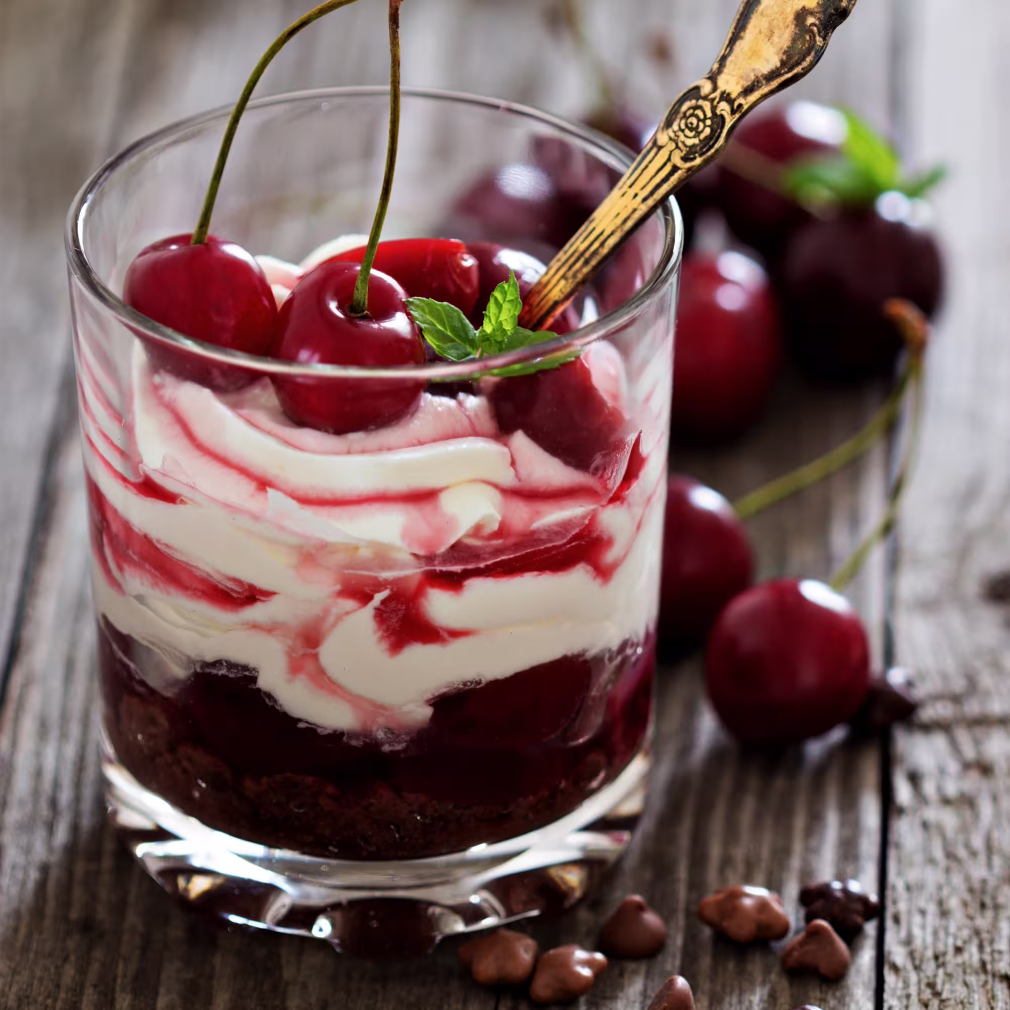 Glass parfait dessert with layers of whipped cream, cherry sauce, fresh cherries, and a mint leaf on top.