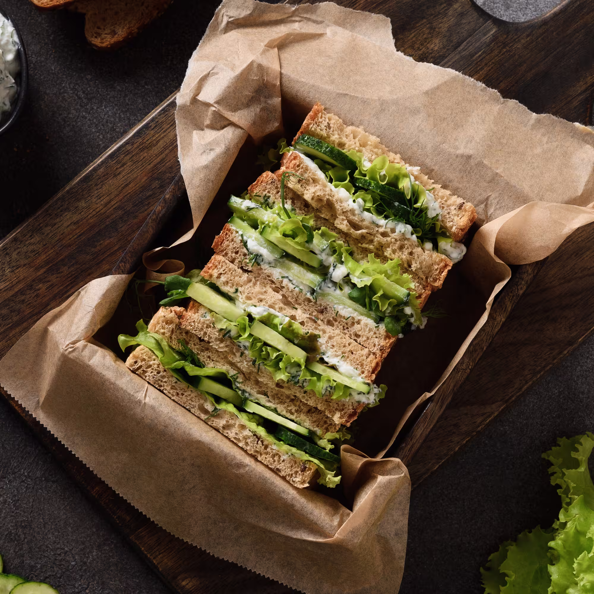 Three cucumber and lettuce tea sandwiches on whole wheat bread, arranged on brown parchment paper in a wooden tray.