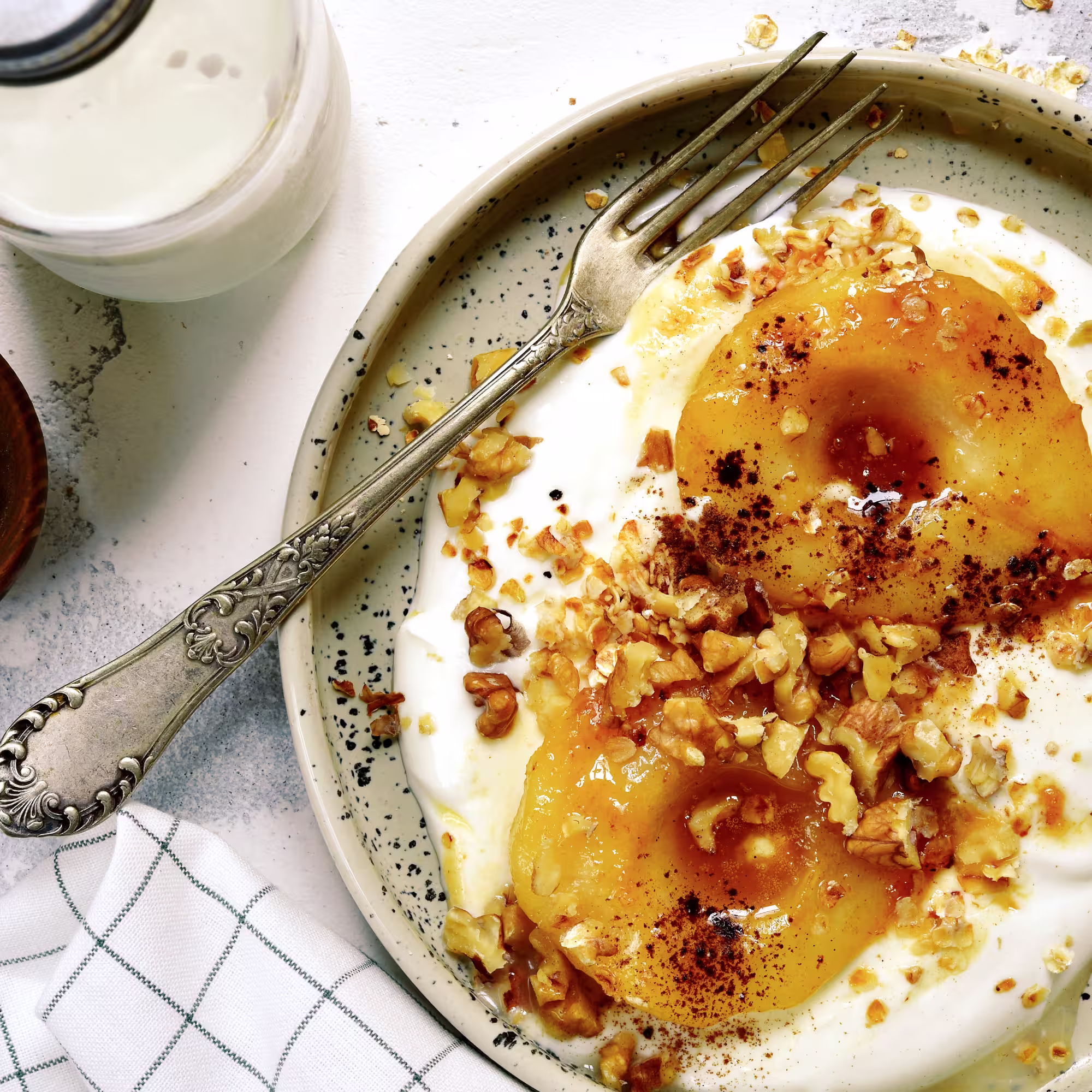 Baked pear halves served on yogurt, topped with chopped walnuts and sprinkled with cinnamon in a speckled bowl with a vintage fork.