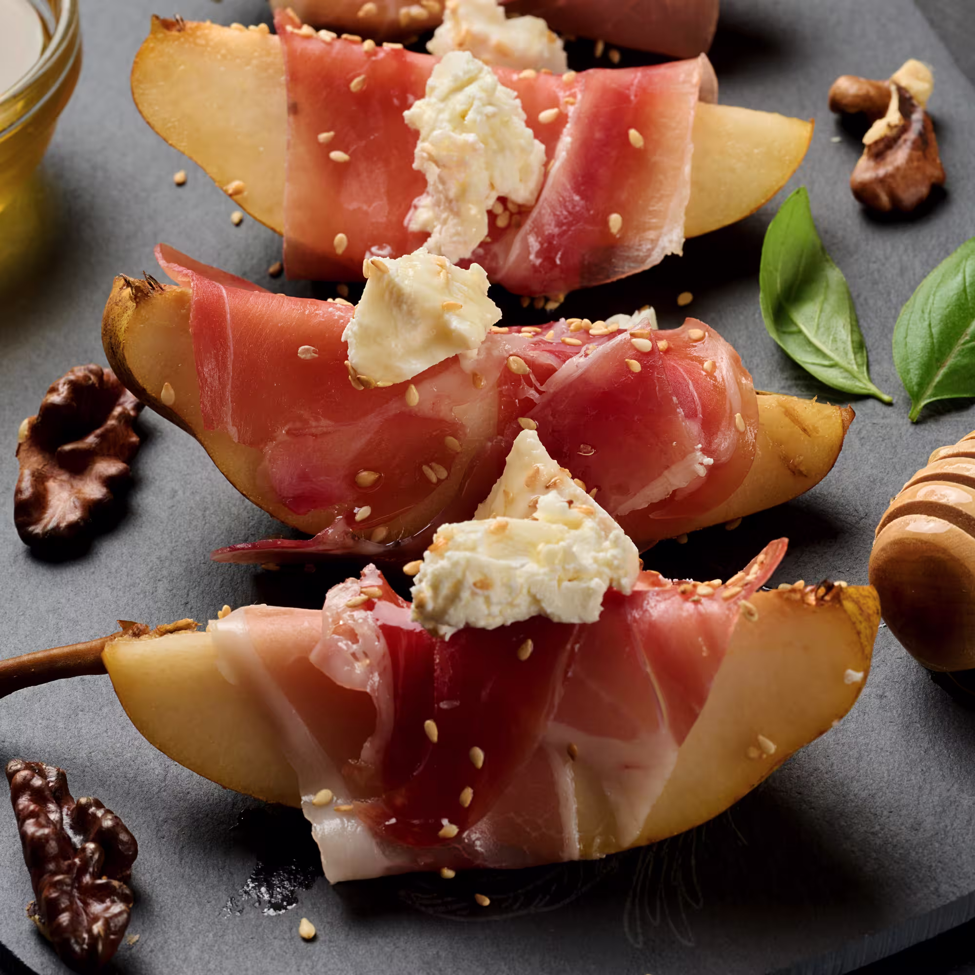 Pear slices wrapped in prosciutto, topped with creamy cheese and sprinkled with sesame seeds on a dark surface with walnuts and basil leaves nearby.