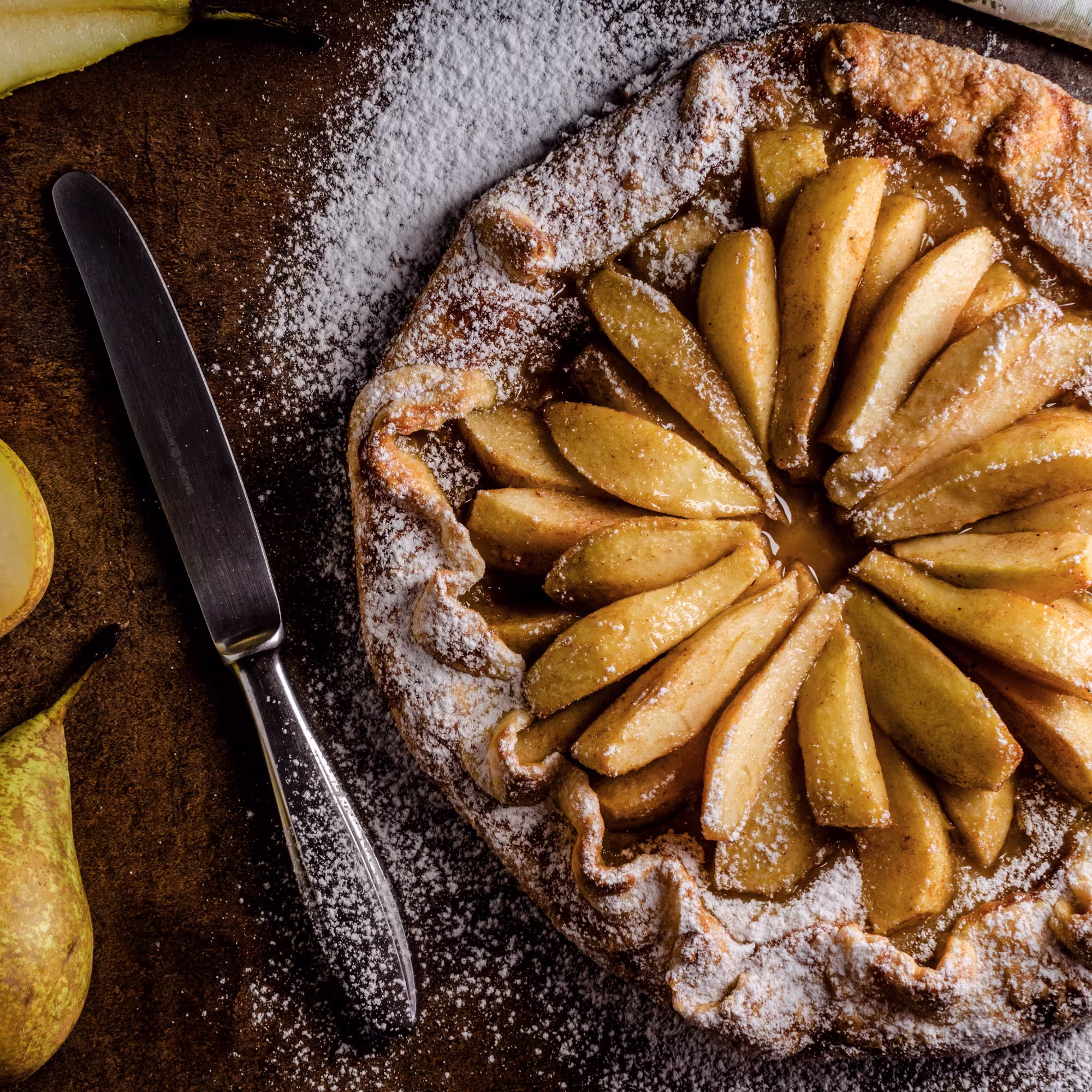 Rustic pear tart dusted with powdered sugar on a dark surface next to a knife and fresh pears.