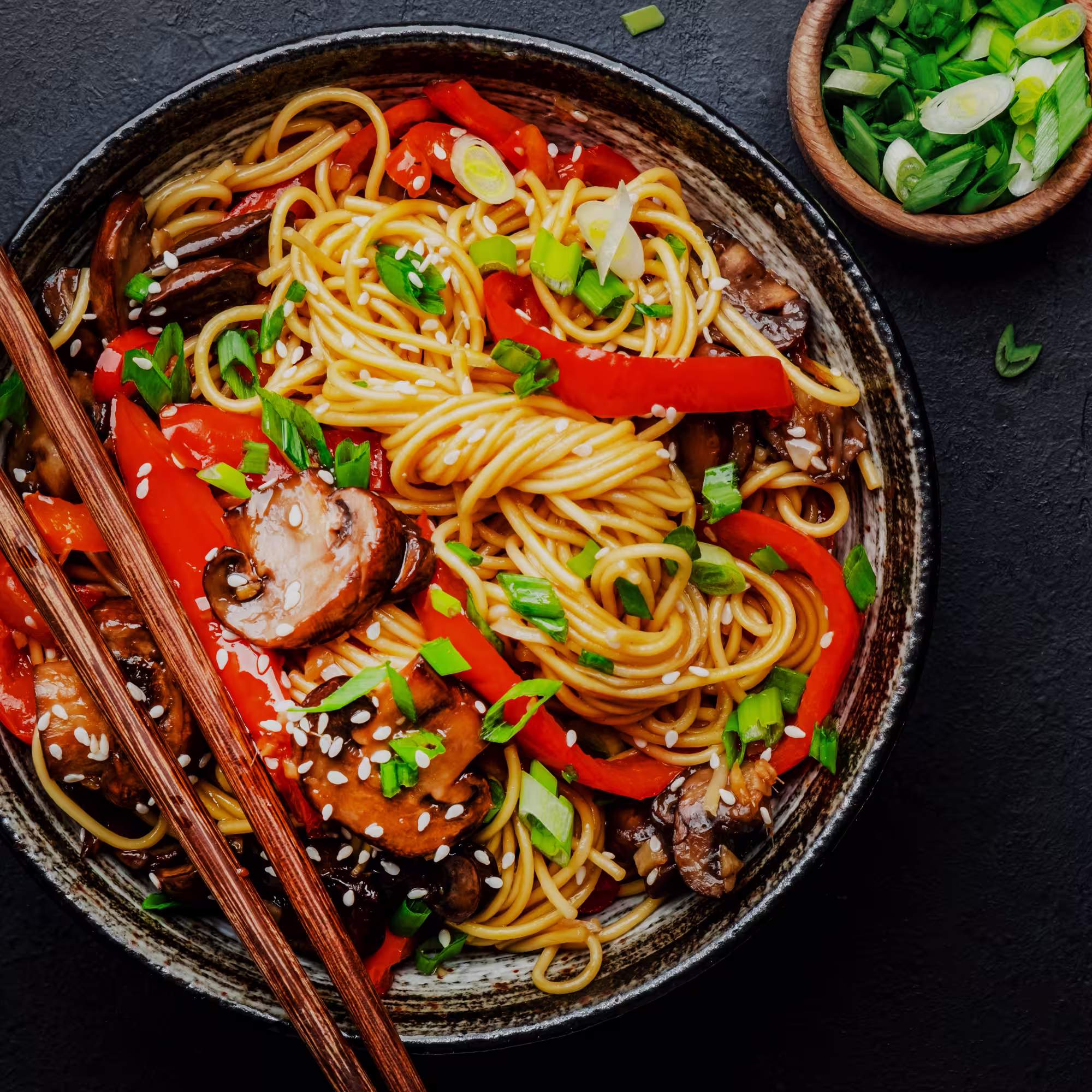 Bowl of stir-fried noodles with mushrooms, red bell peppers, green onions, and sesame seeds with wooden chopsticks.