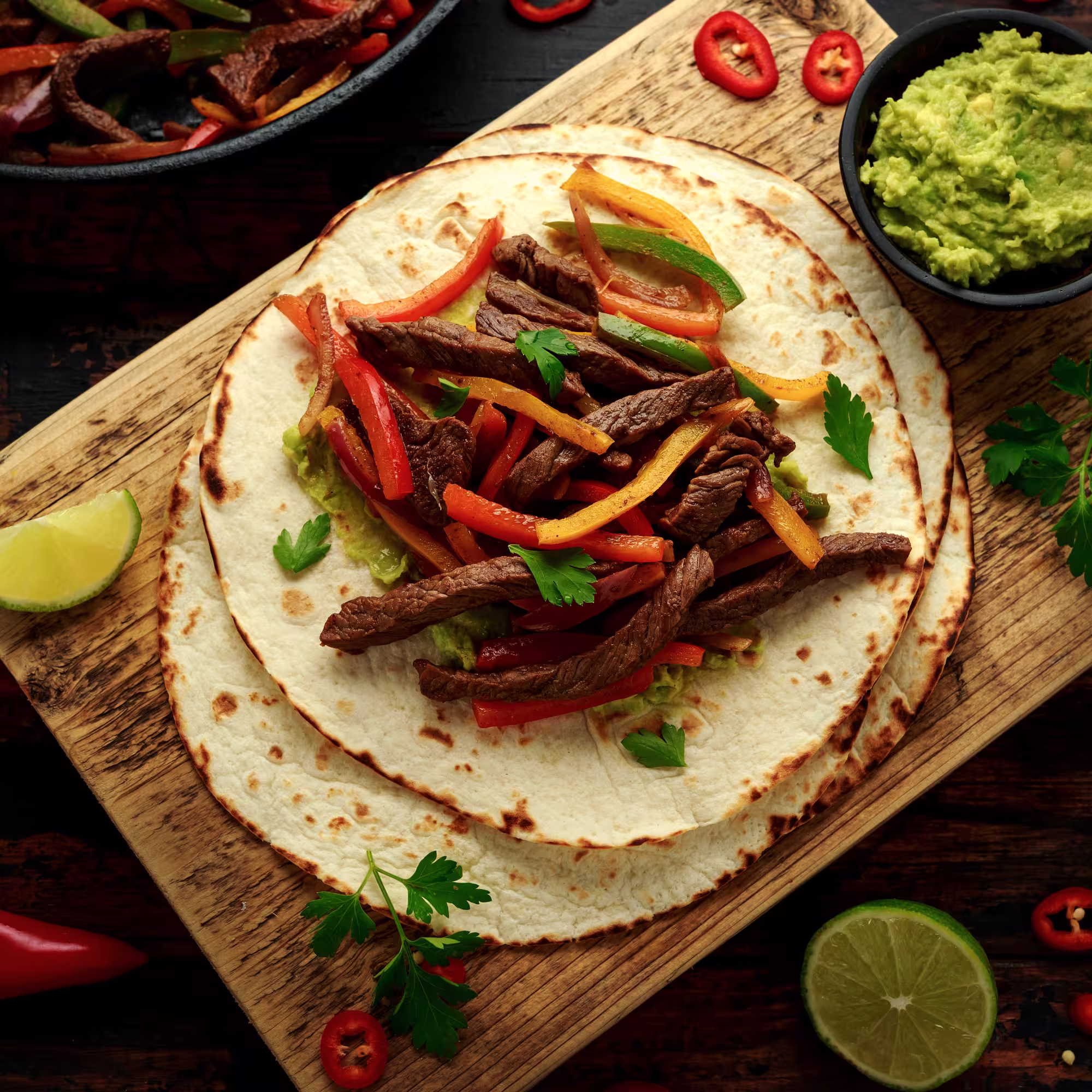 Flour tortilla topped with grilled steak strips, colorful bell peppers, and guacamole on a wooden board with lime wedges and red chili slices nearby.