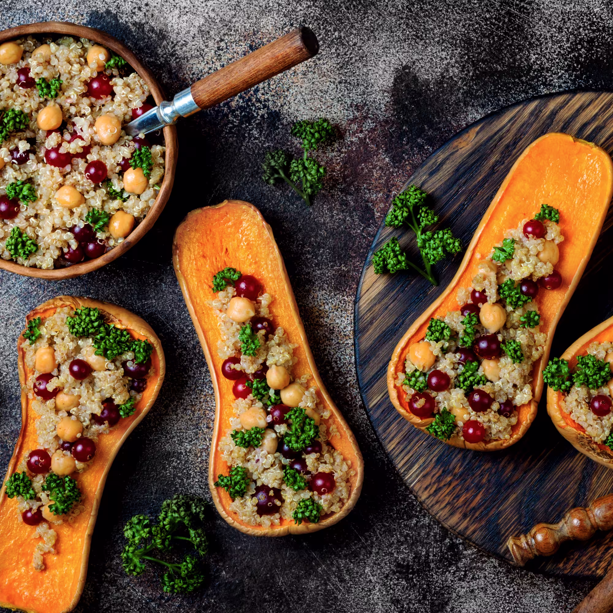 Roasted butternut squash halves filled with quinoa, chickpeas, cranberries, and parsley on a dark textured surface and wooden board.