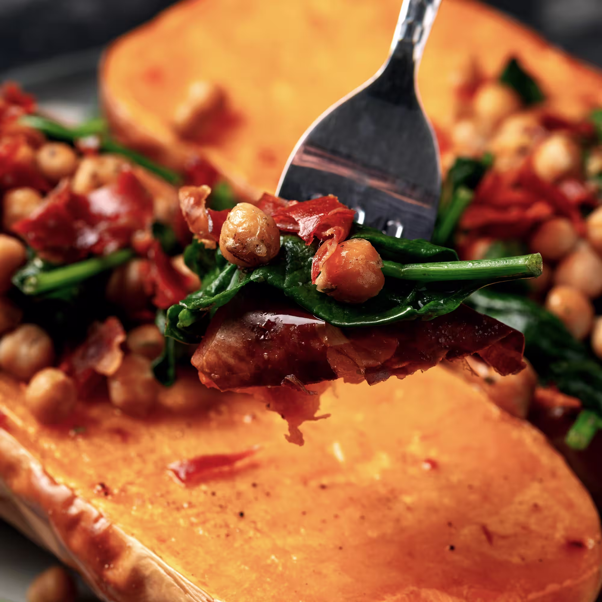 Fork holding roasted chickpeas, wilted spinach, and crispy bacon over a halved roasted butternut squash.