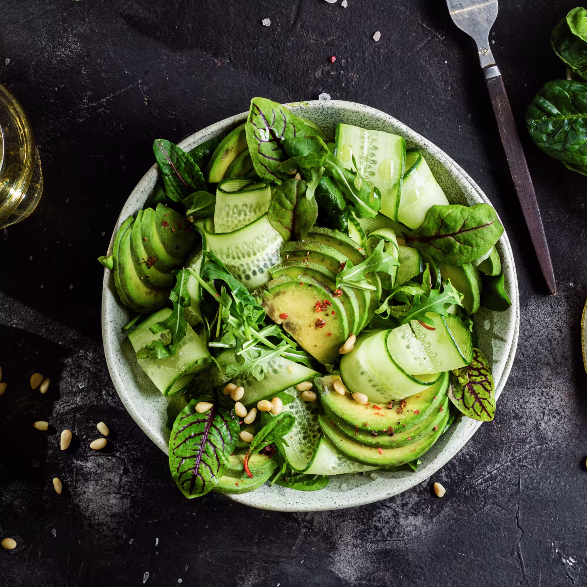 Bowl of fresh green salad with sliced cucumber ribbons, avocado slices, mixed greens, pine nuts, and seasoning on a dark surface.