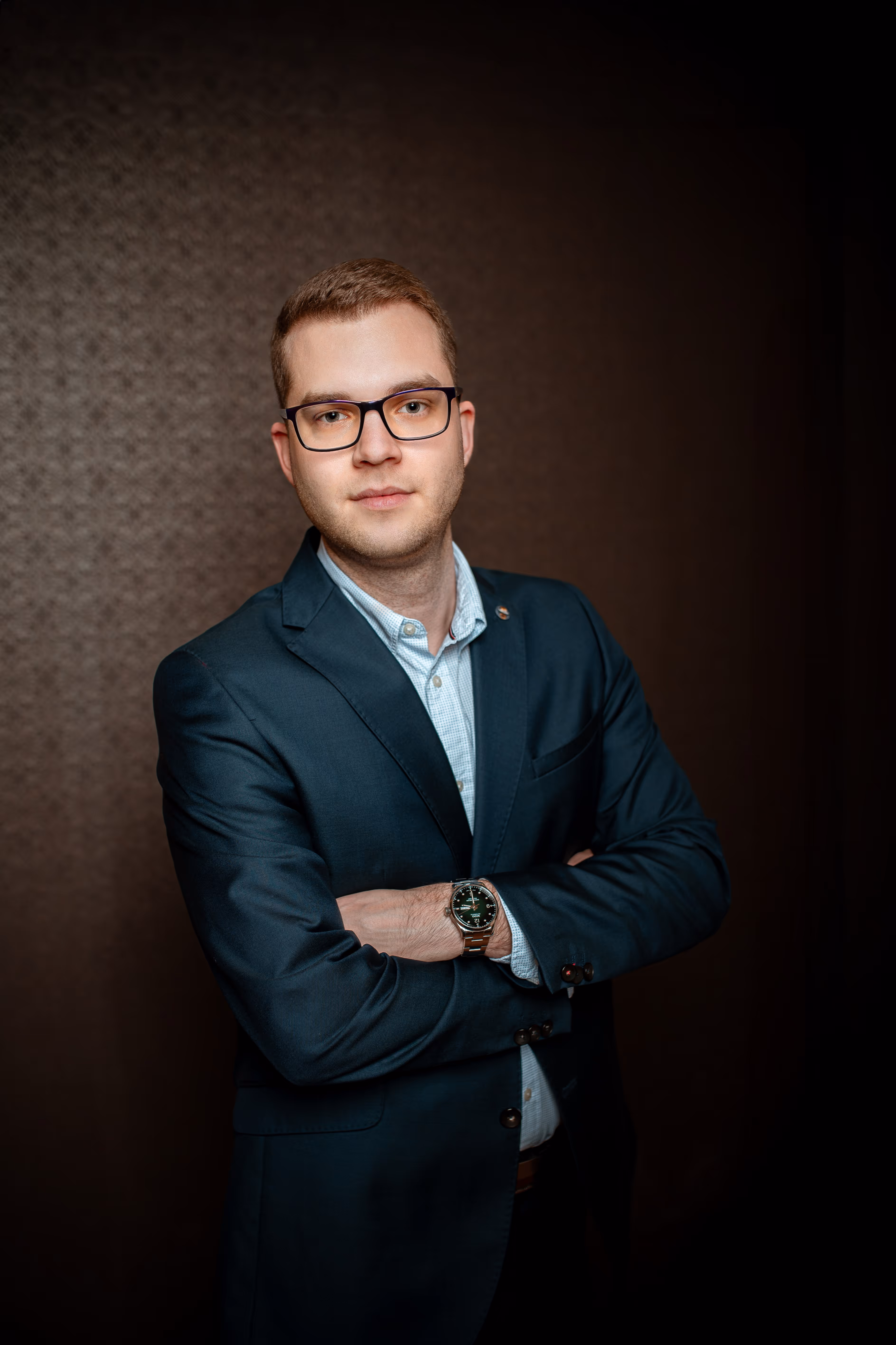 Szakáll nélküli, szemüveges fiatal férfi sötétkék zakóban és világoskék ingben, karját keresztbe téve, barna háttér előtt.