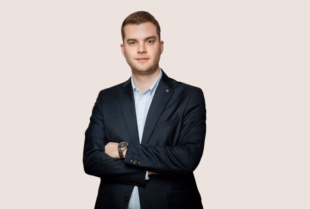 Fiatal férfi sötét zakóban, karját keresztbe téve, bézs háttér előtt.