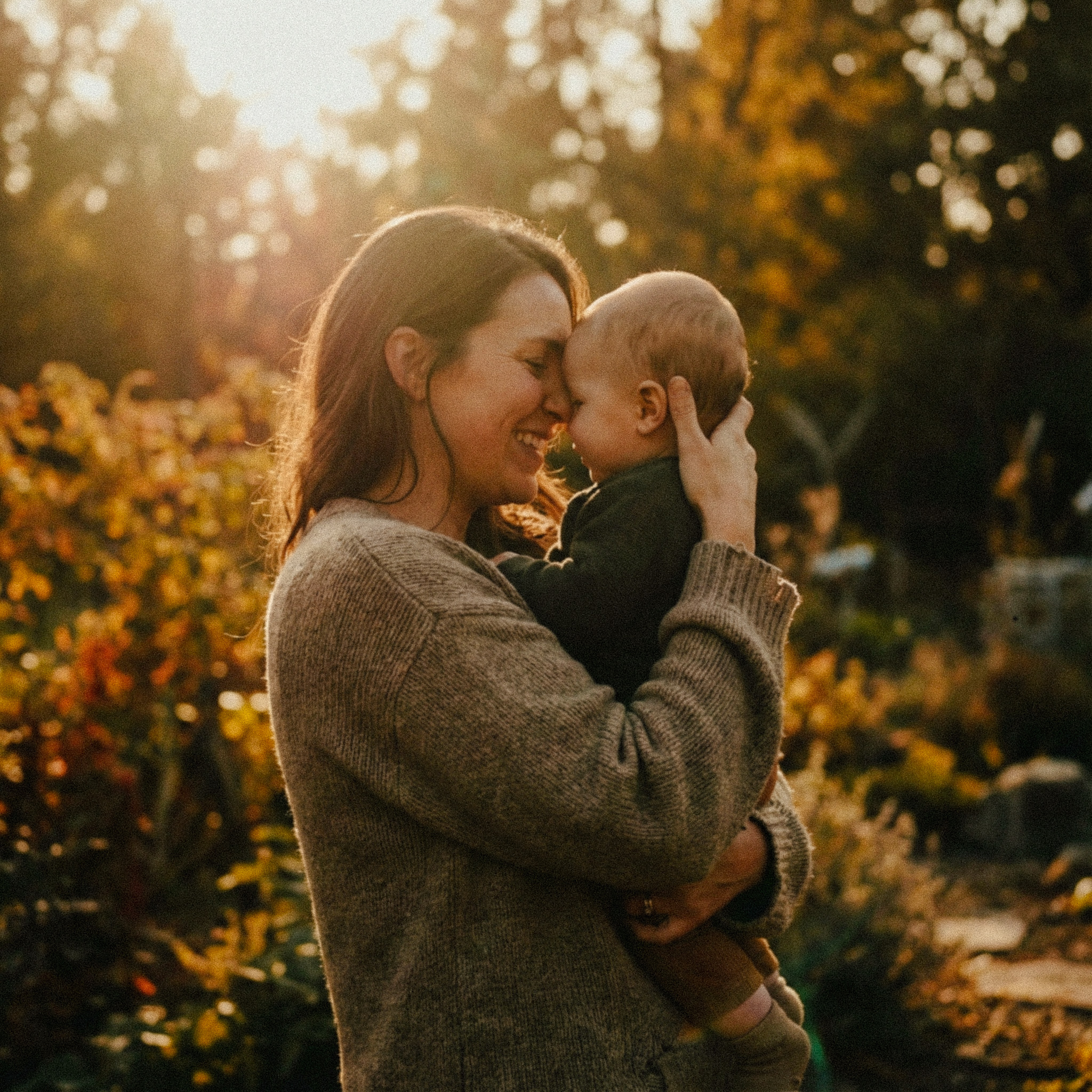 Woman in a brown sweater holding and smiling with a baby outdoors in a sunlit garden.