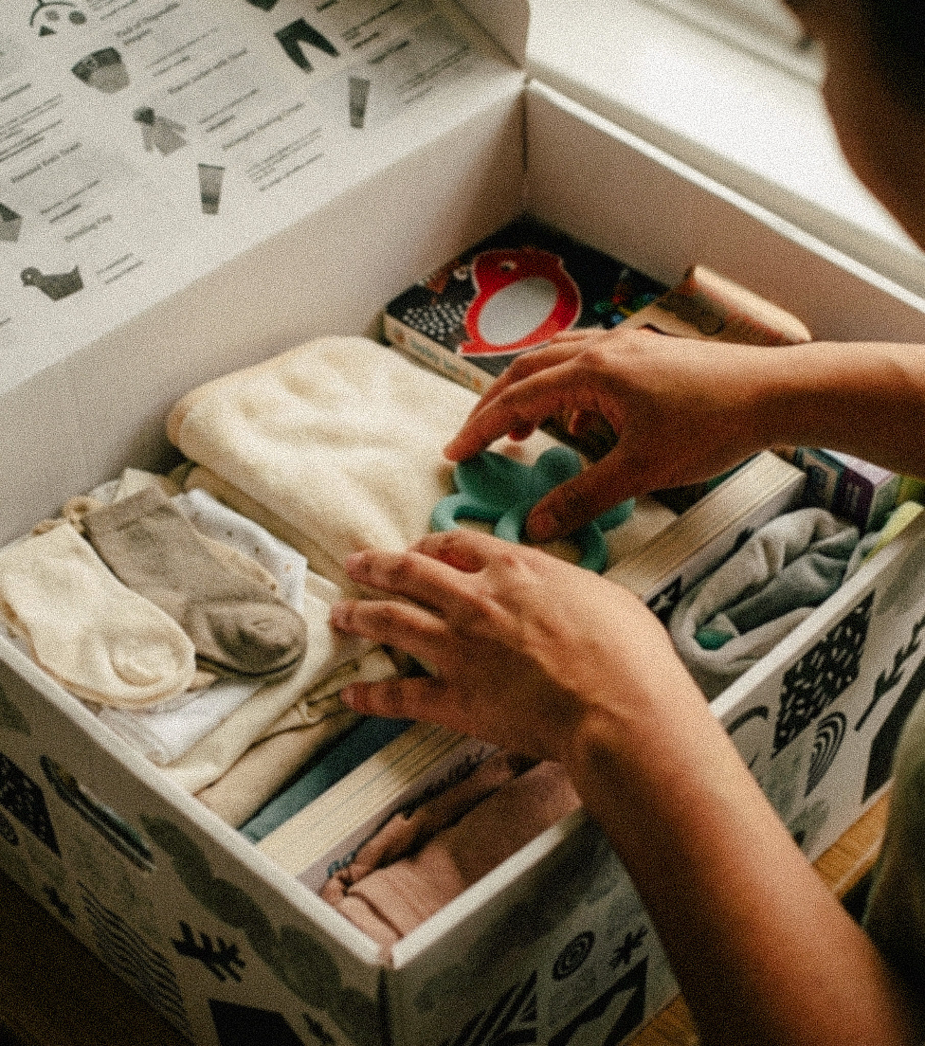 Hands organizing folded baby clothes, socks, and a green toy inside a decorated baby bundle box.
