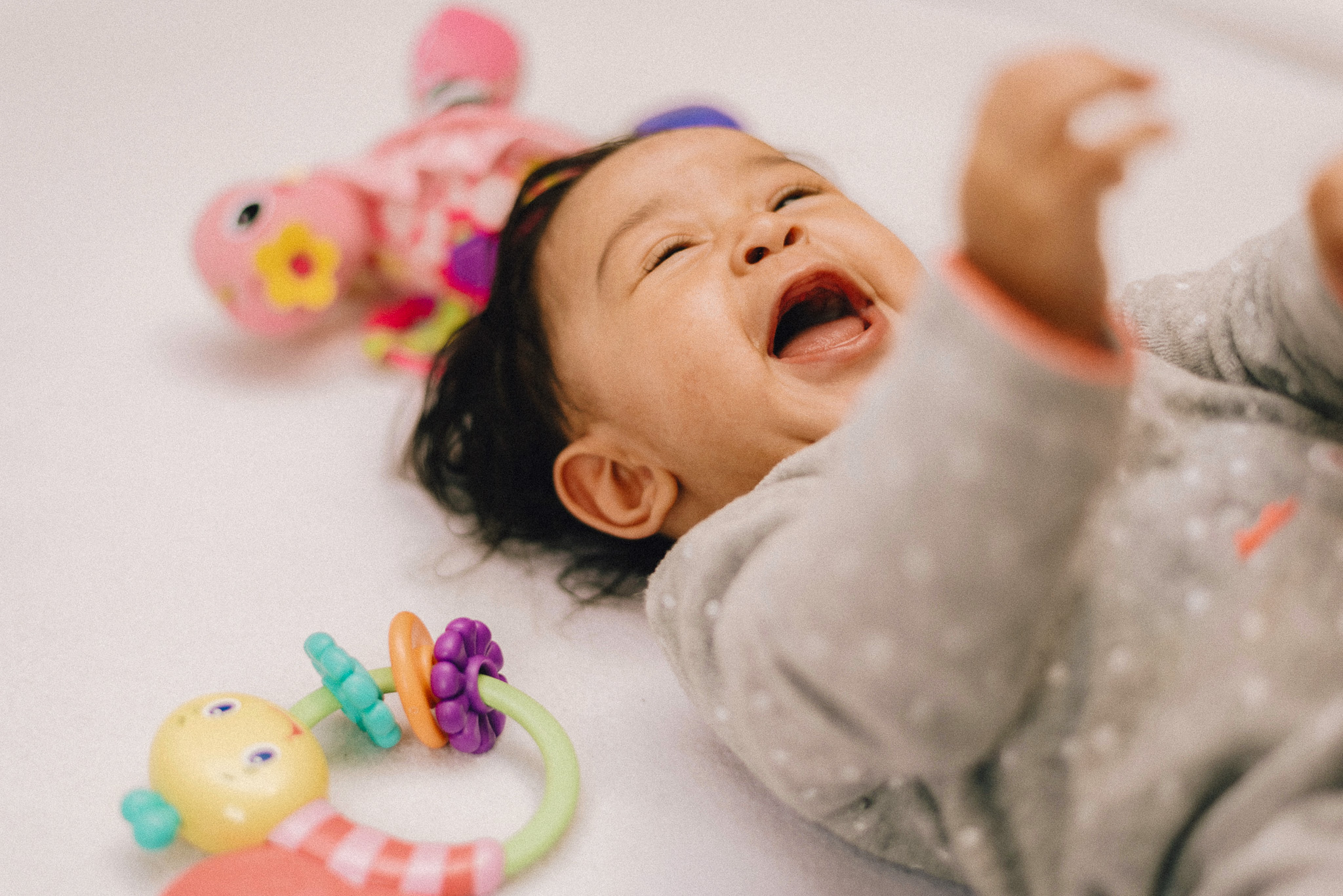 Laughing baby lying on a white surface with colorful toys nearby.