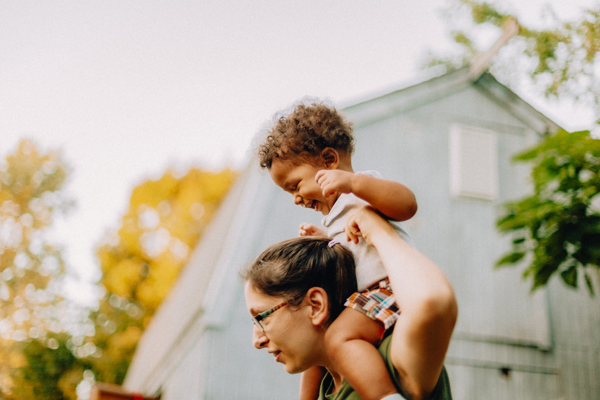 Adult carrying a smiling toddler on their shoulders outdoors near a blue building with trees in the background.