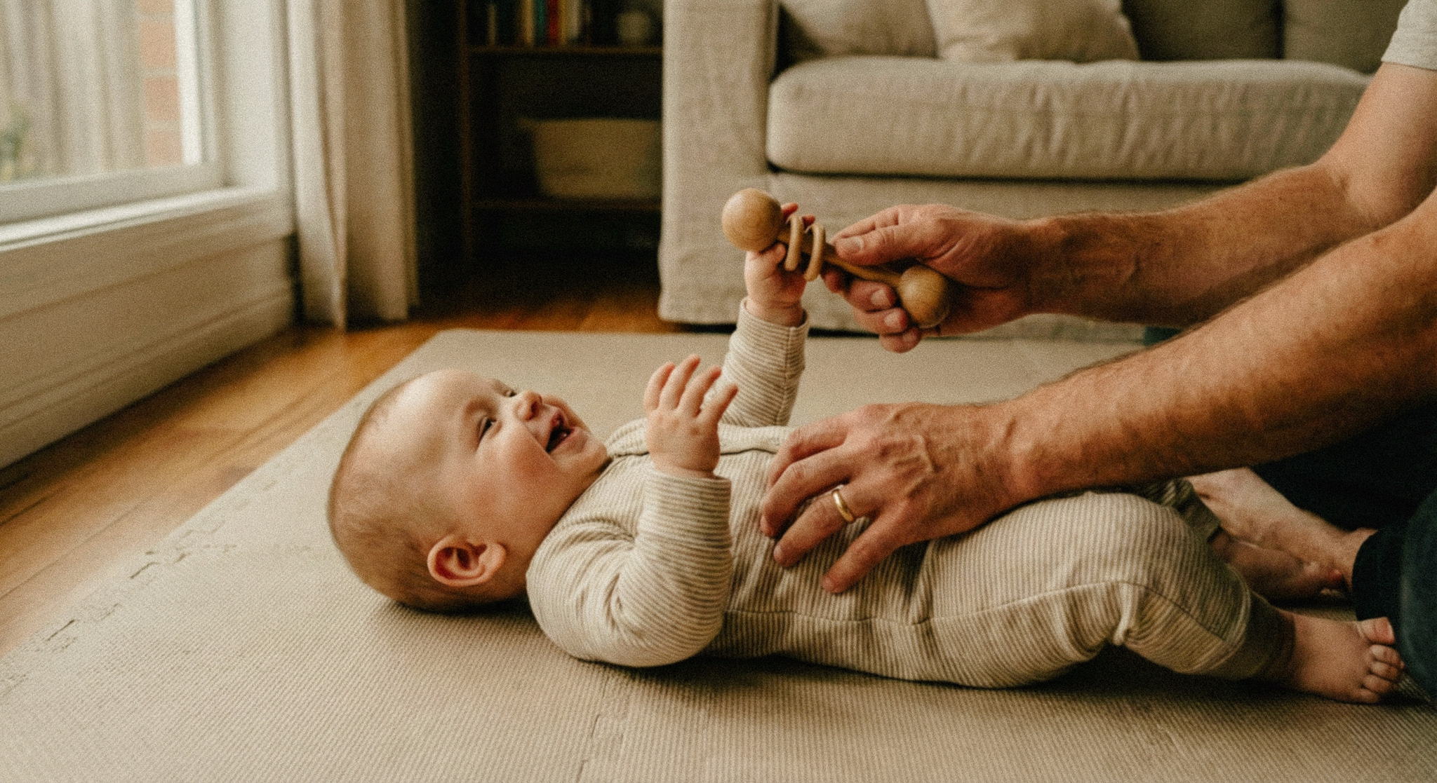 Smiling baby lying on a soft mat, reaching for a wooden rattle held by an adult.