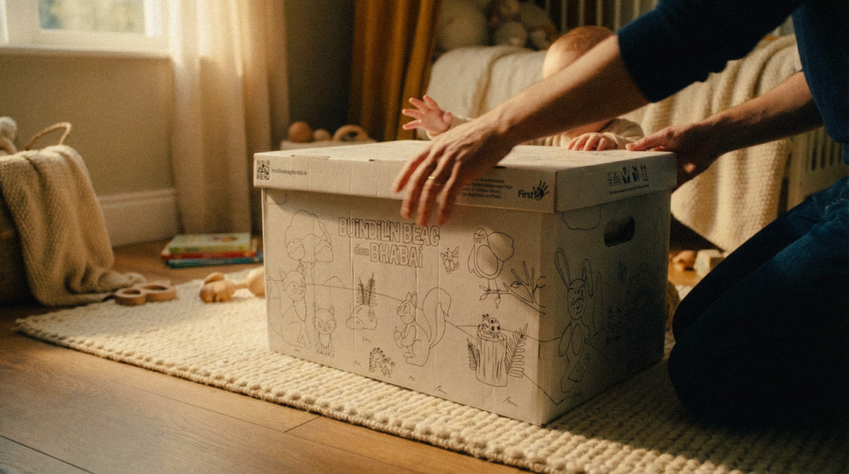 Hands opening a large cardboard box decorated with animal sketches on a carpeted floor, with a baby reaching over the box in a softly lit room.