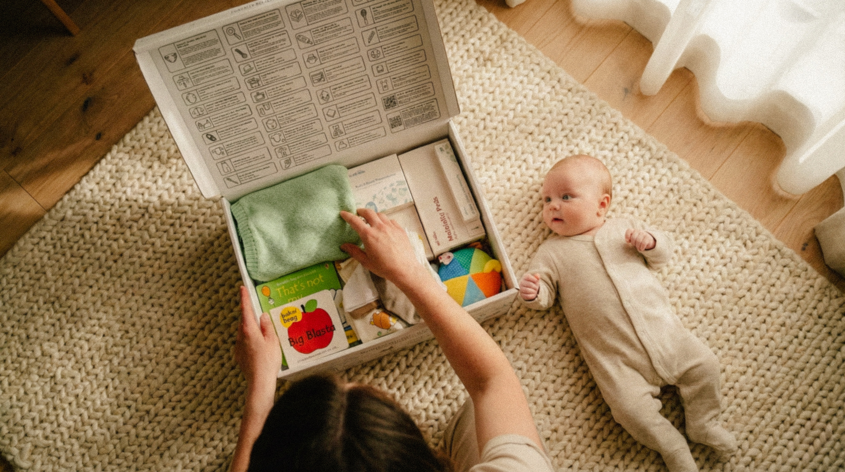 Top view of a woman opening a baby care box next to a baby in a beige onesie lying on a knitted cream rug.