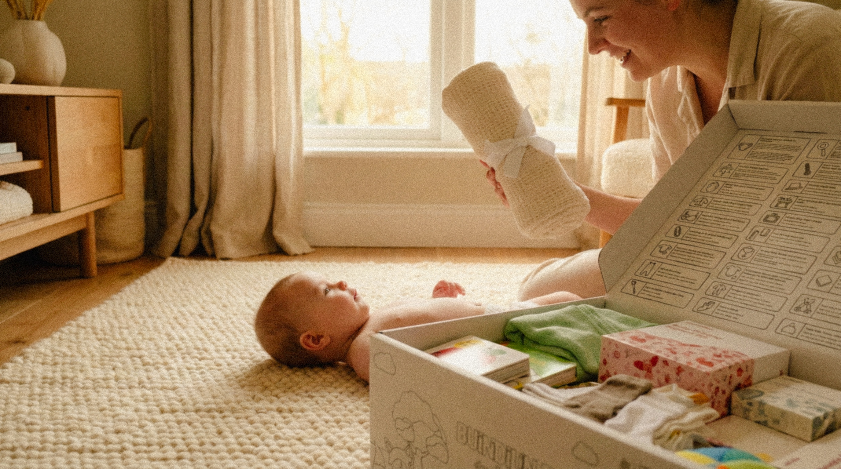 Smiling woman holding a rolled blanket while a baby lies on a textured cream rug near an open box of baby items.