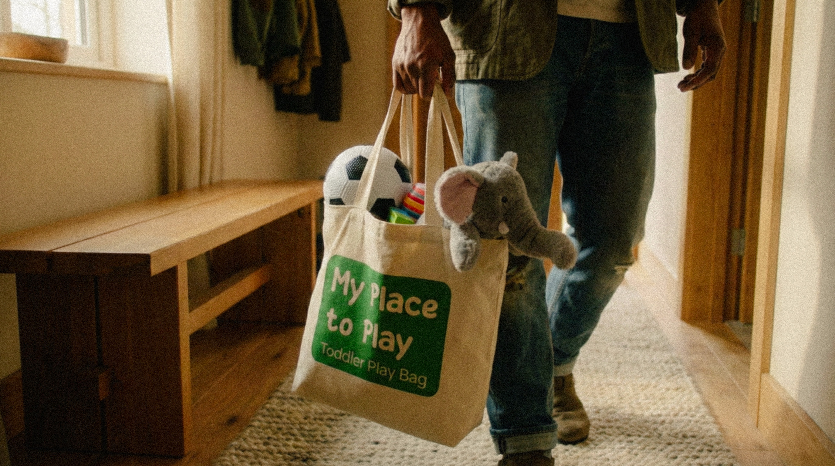 Person holding a canvas tote bag labeled 'My Place to Play Toddler Play Bag' filled with a soccer ball, colorful toys, and a stuffed elephant.