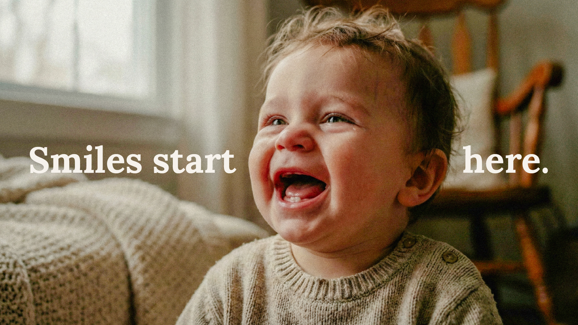 Baby wearing a beige sweater laughing joyfully indoors with the text 'Smiles start here.' overlaid.