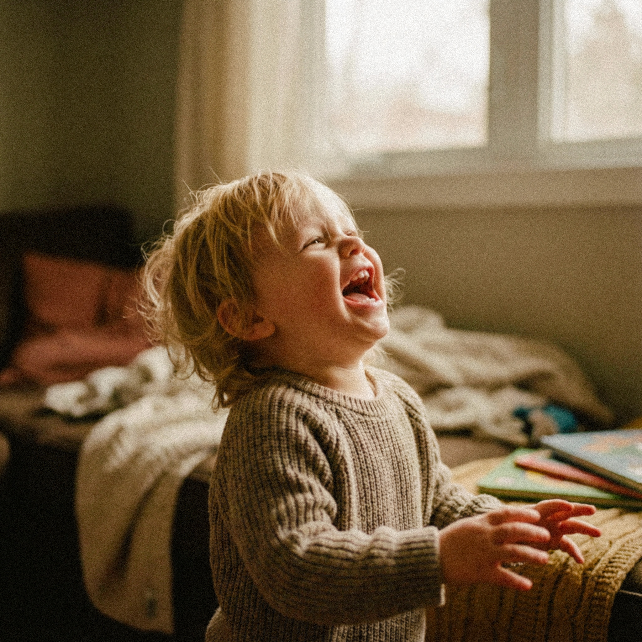 Toddler with blonde hair laughing joyfully indoors near a window.