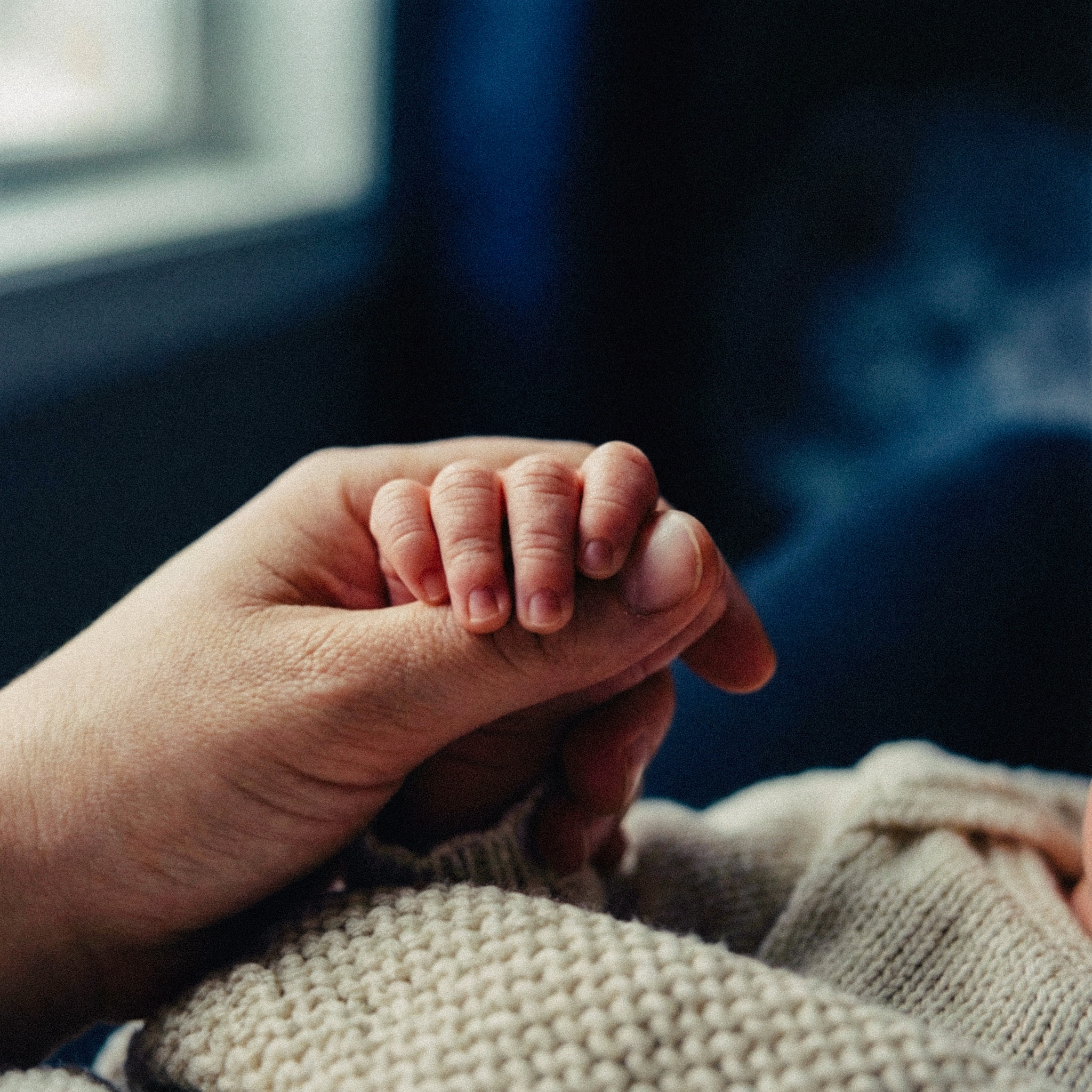 Close-up of an adult hand gently holding a baby's hand wrapped in a knitted blanket.