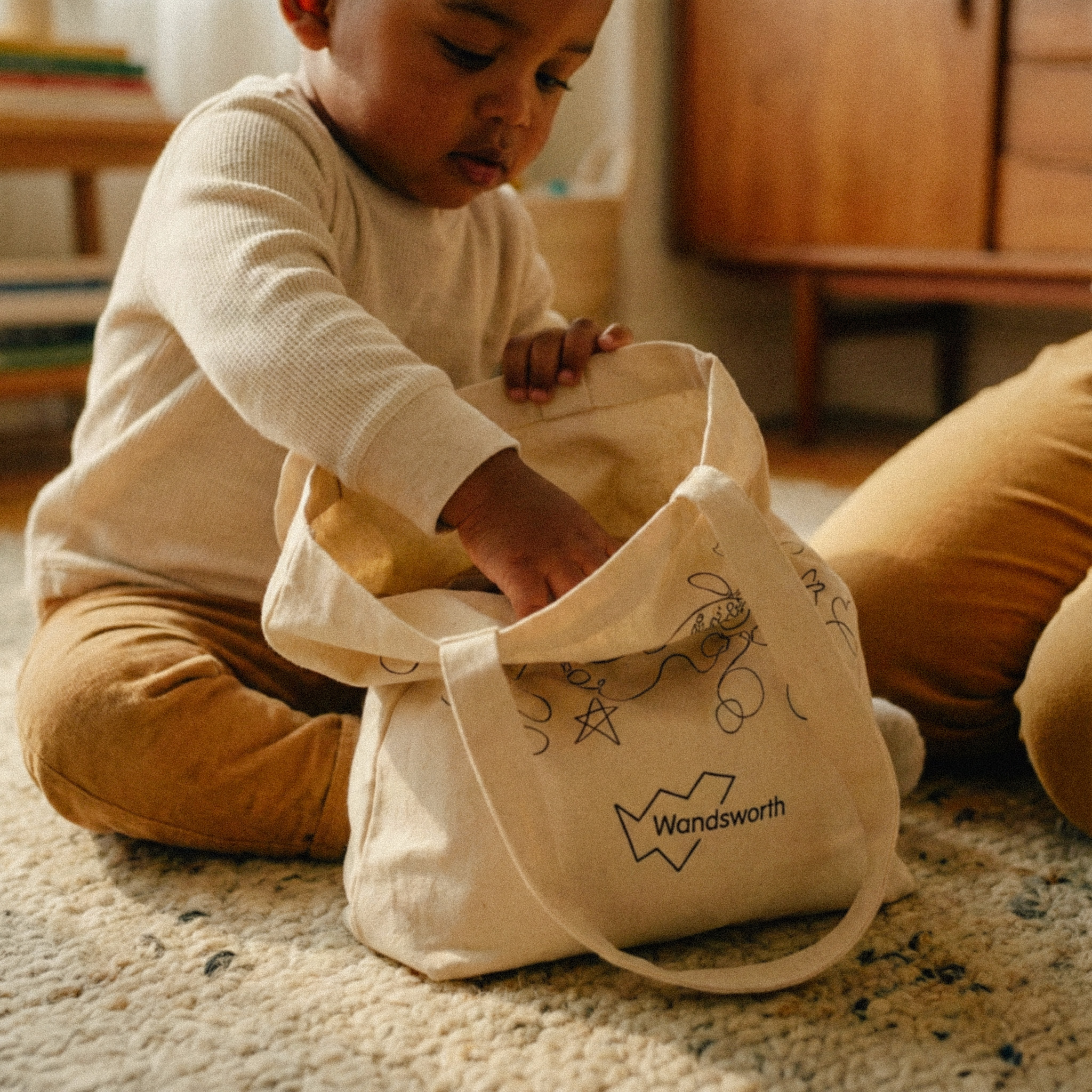 Child sitting on a carpeted floor reaching into a beige tote bag labeled 'Wandsworth' with doodle designs.