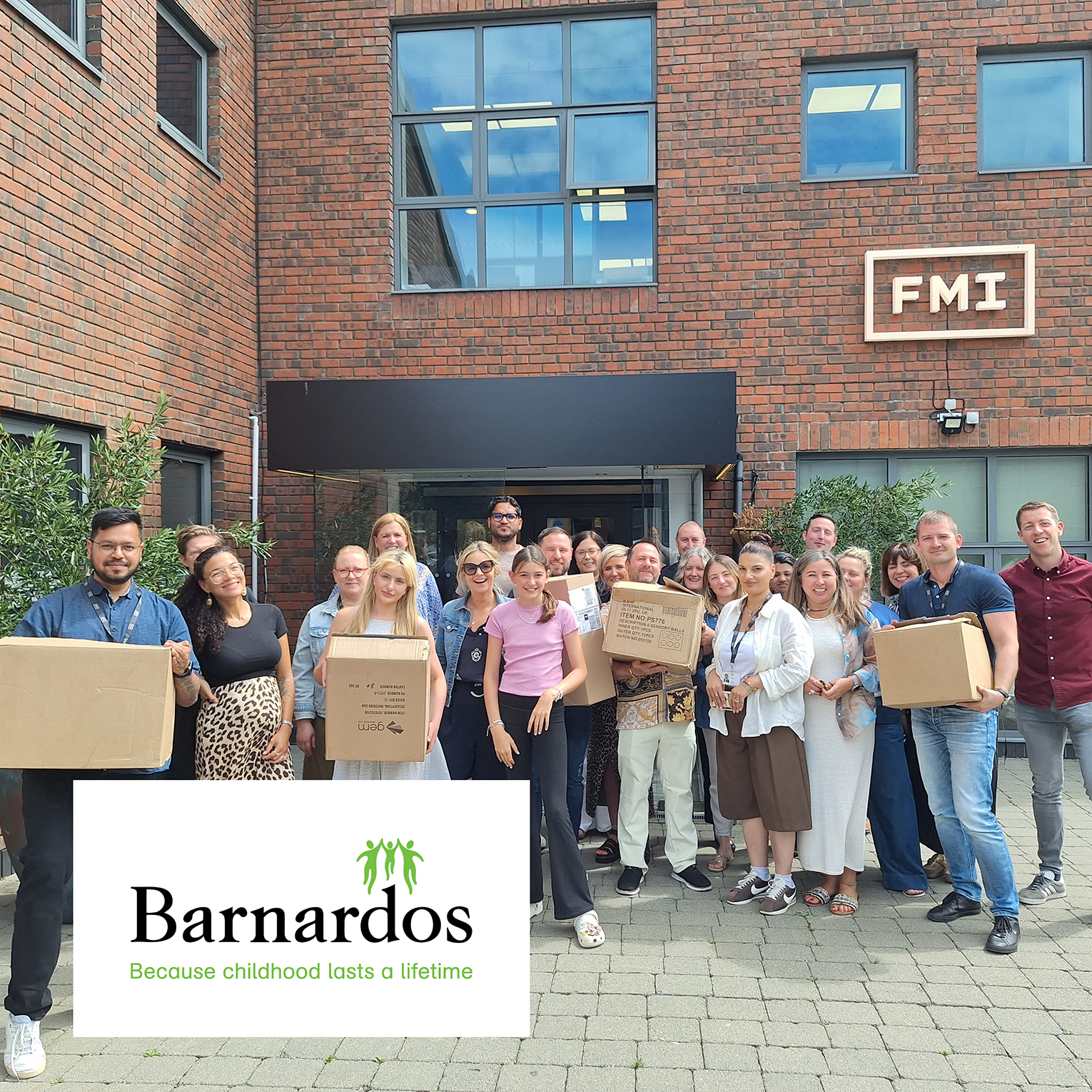Group of diverse adults standing outside a building with bricks, holding boxes and smiling, with Barnardos logo and tagline about childhood.