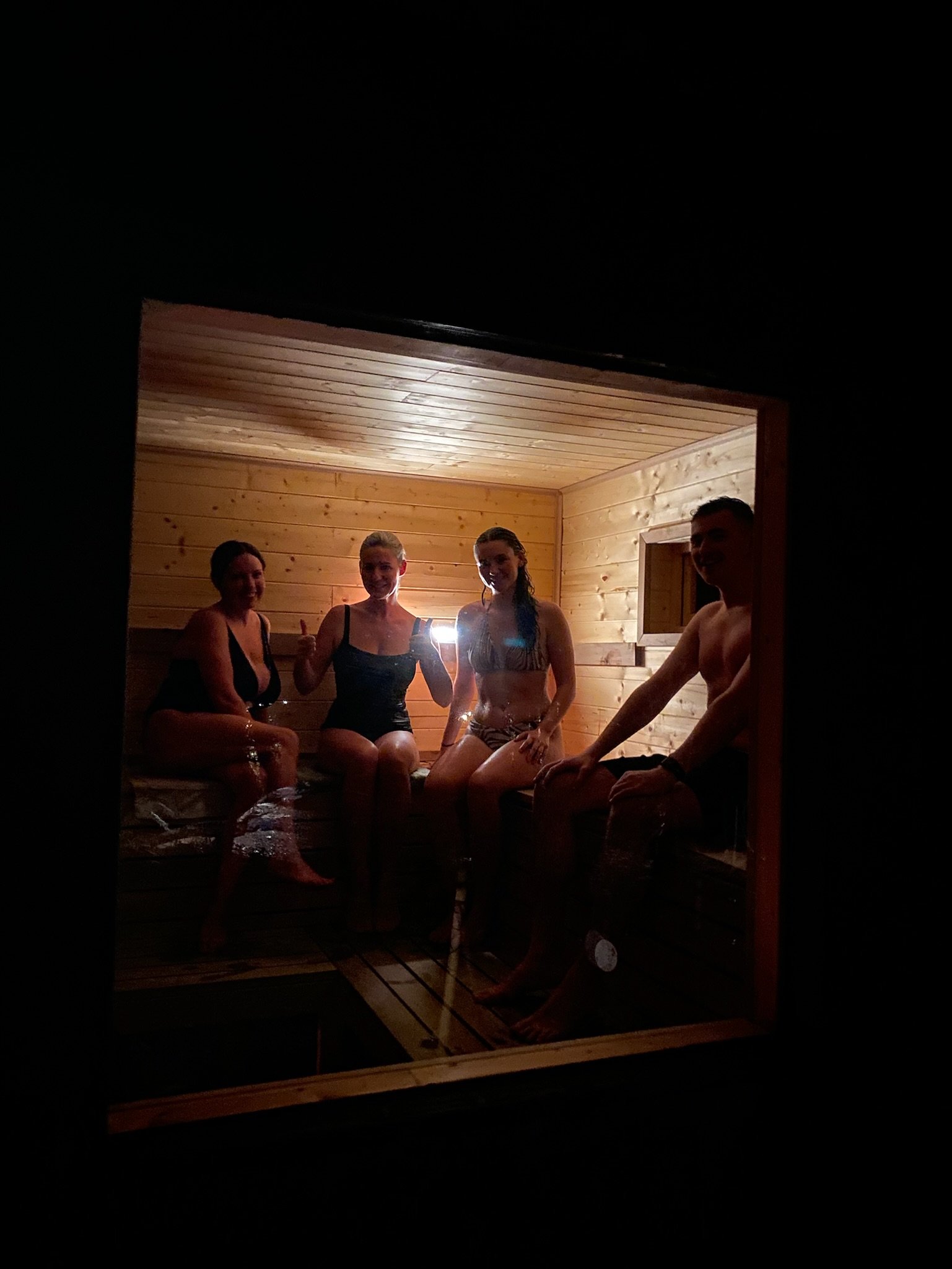 Four people sitting inside a wooden sauna room, wearing swimwear, visible through a glass window with dim lighting.