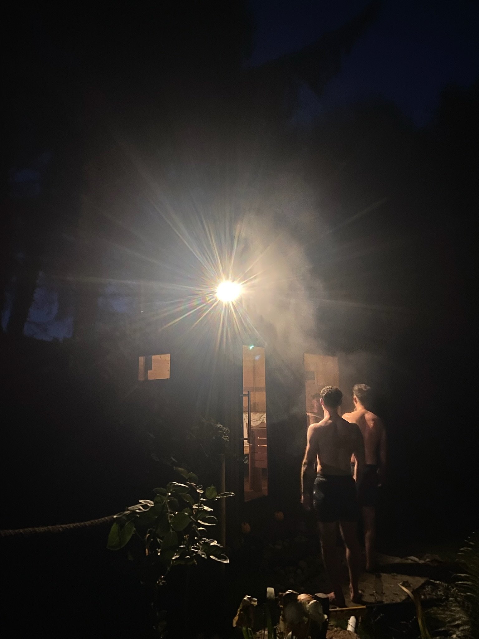 Two shirtless men standing outside a dimly lit building at night with smoke or mist illuminated by a bright light above the entrance.