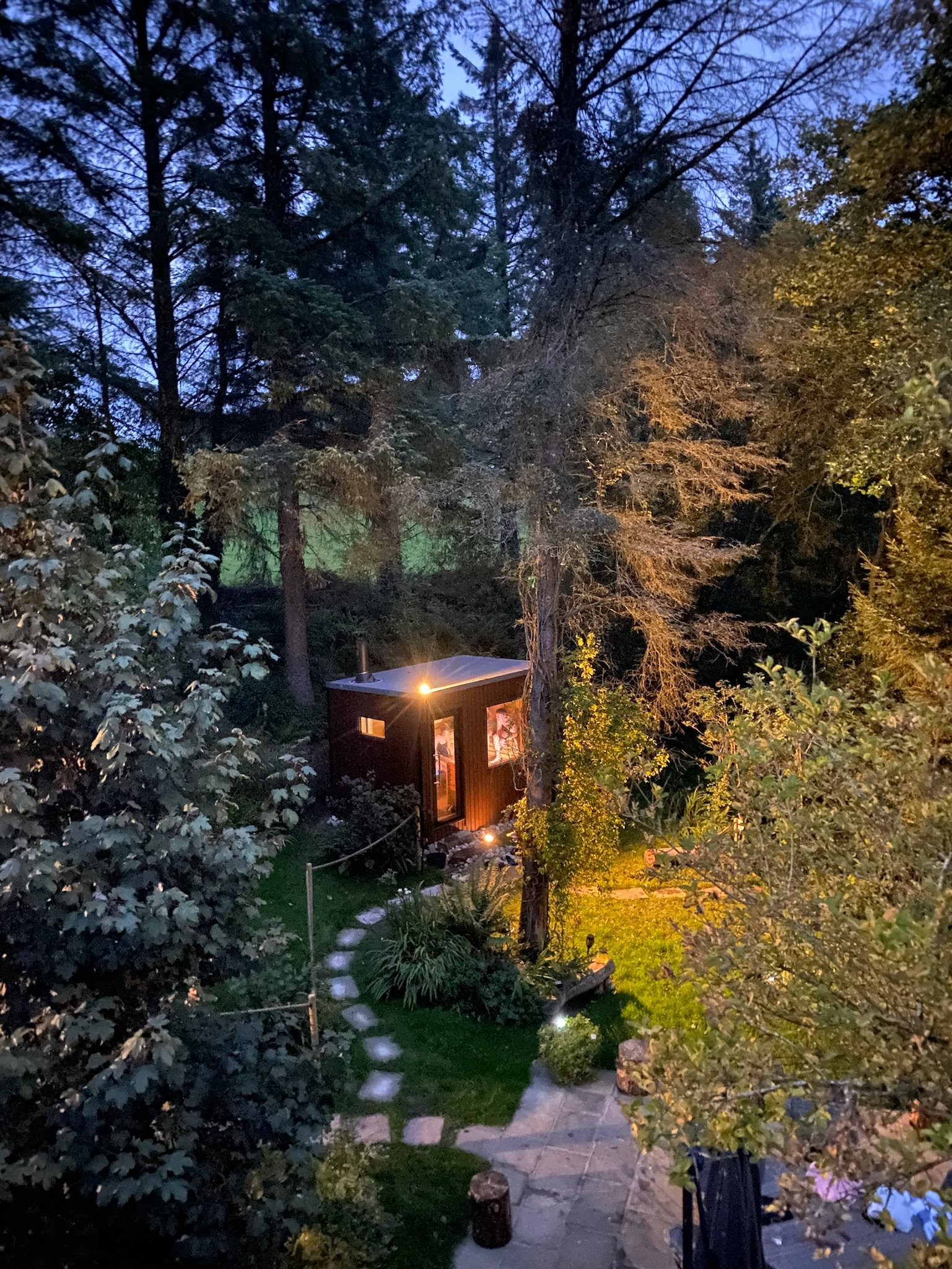 Small illuminated cabin surrounded by trees in a backyard garden during twilight.