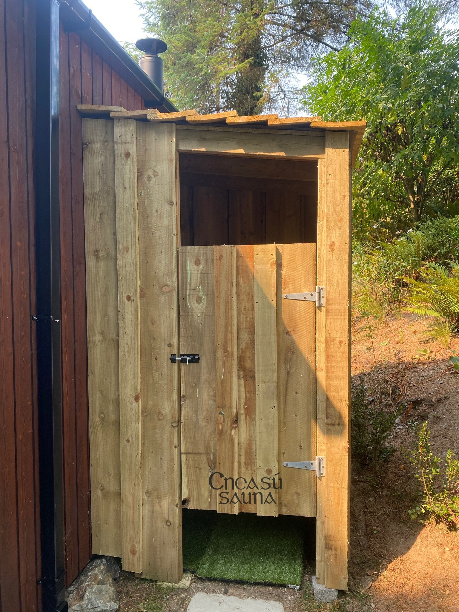 Small wooden outdoor sauna structure with a half door, labeled 'Cneasú Sauna', next to a brown building and surrounded by greenery.