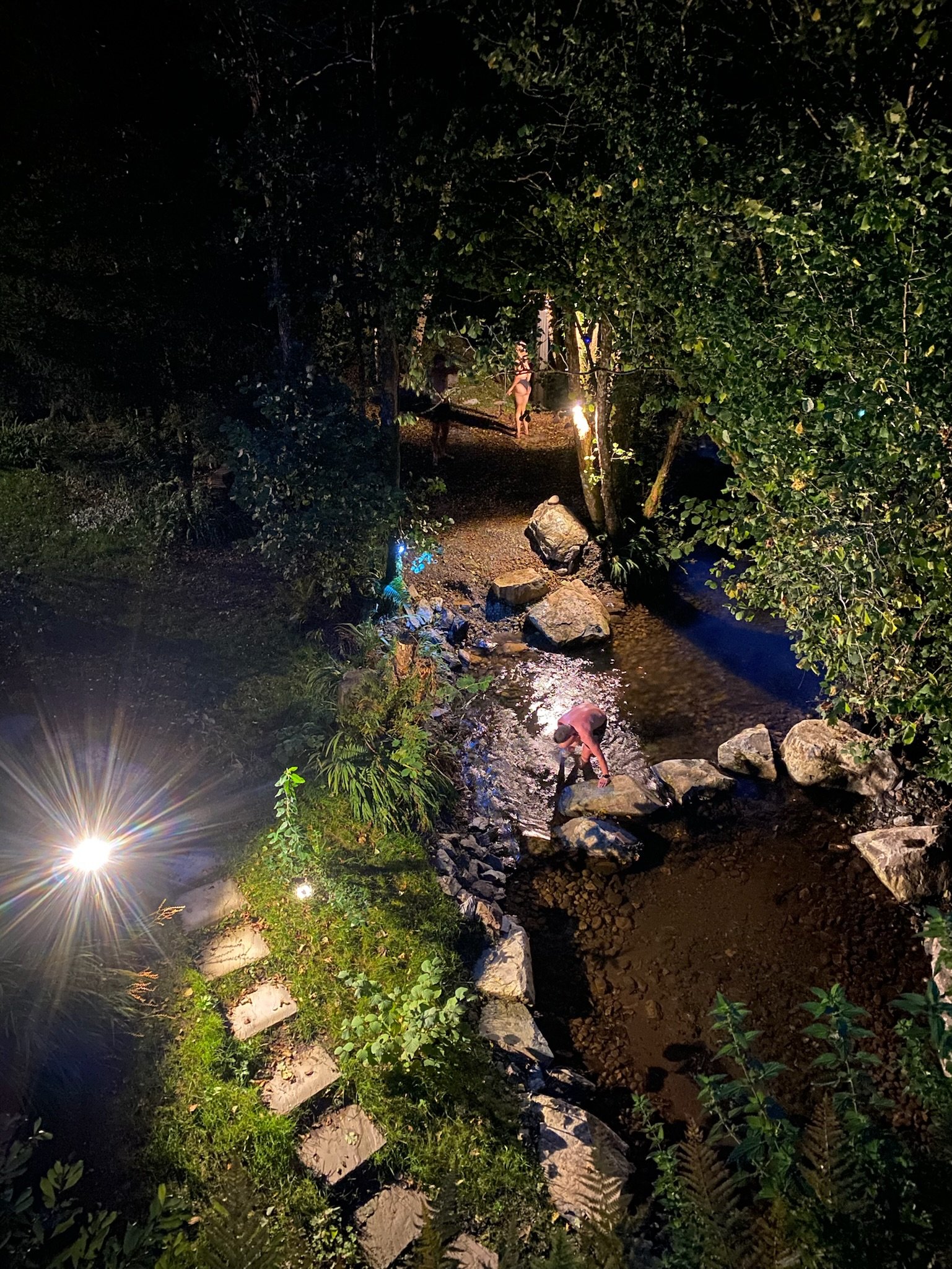 Night scene of a small creek with rocks and stepping stones lit by bright lights, showing a person bent over near the water and two people walking along the path in the background.