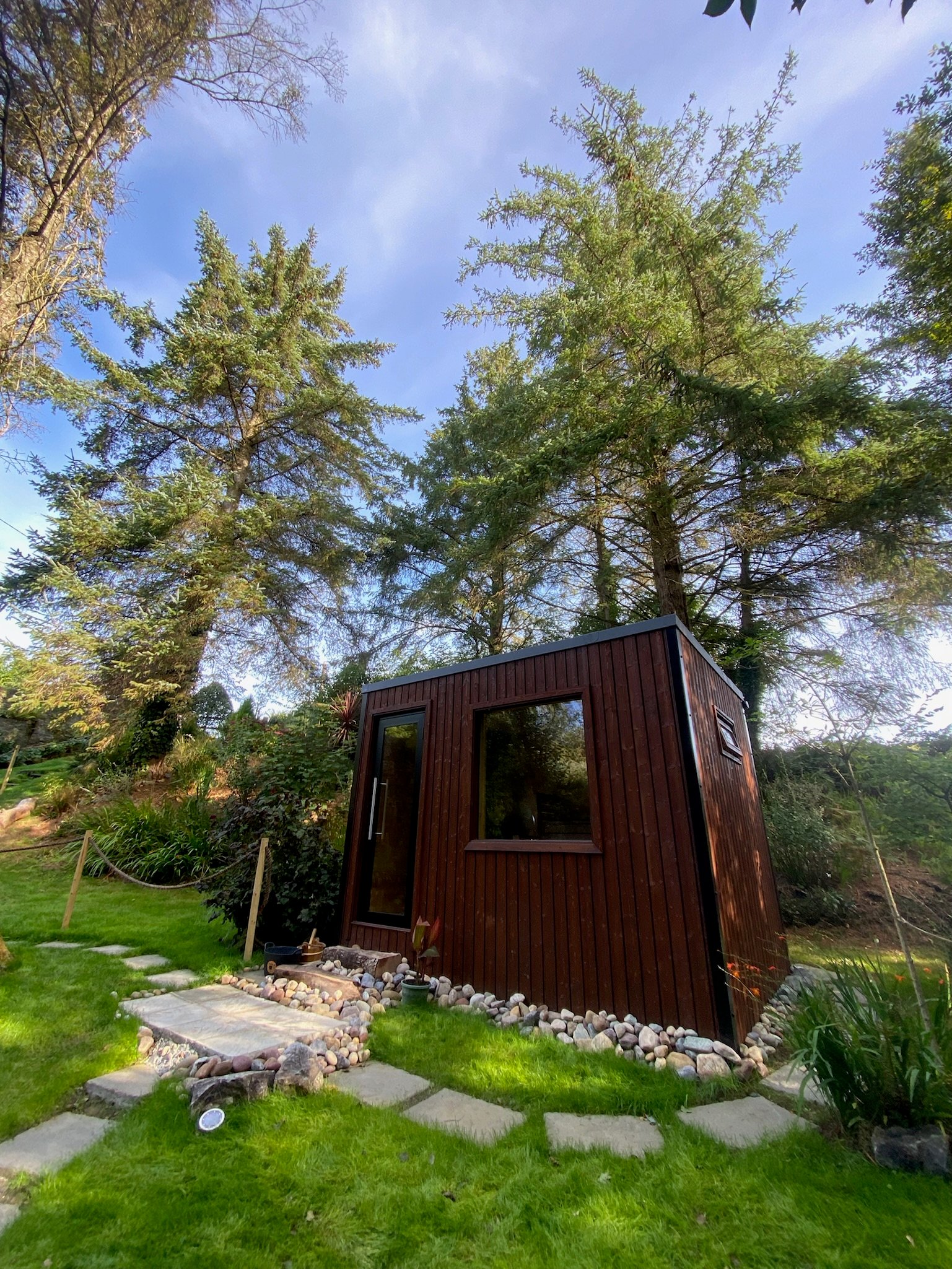 Small modern wooden cabin surrounded by green grass, stone path, and tall evergreen trees under a blue sky.