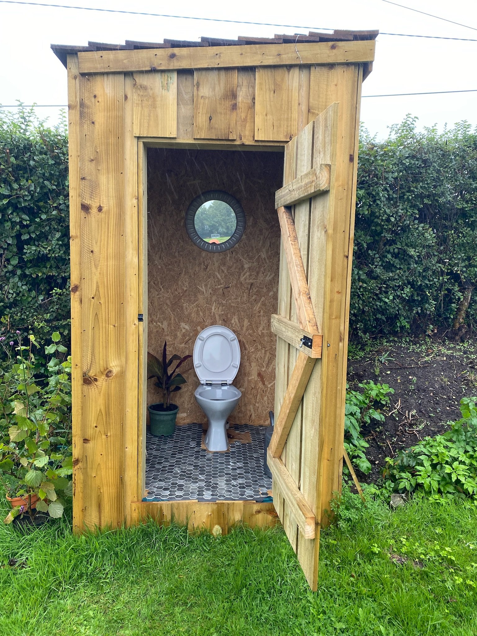 Small wooden outdoor toilet with an open door showing a white toilet, a potted plant, and a circular window.