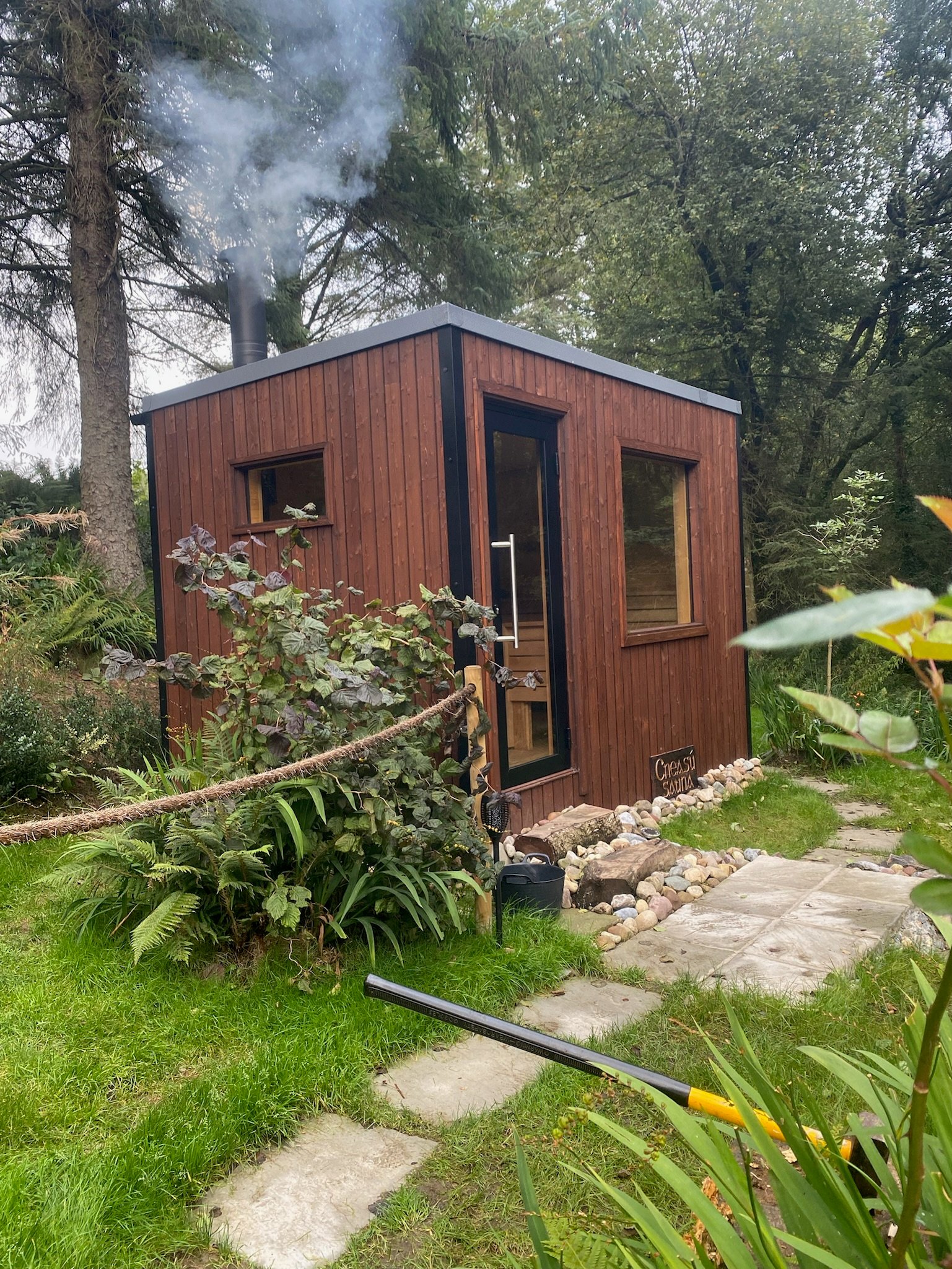 Small wooden sauna cabin with a glass door emitting smoke from a chimney, surrounded by green plants and trees with a stone path leading to it.