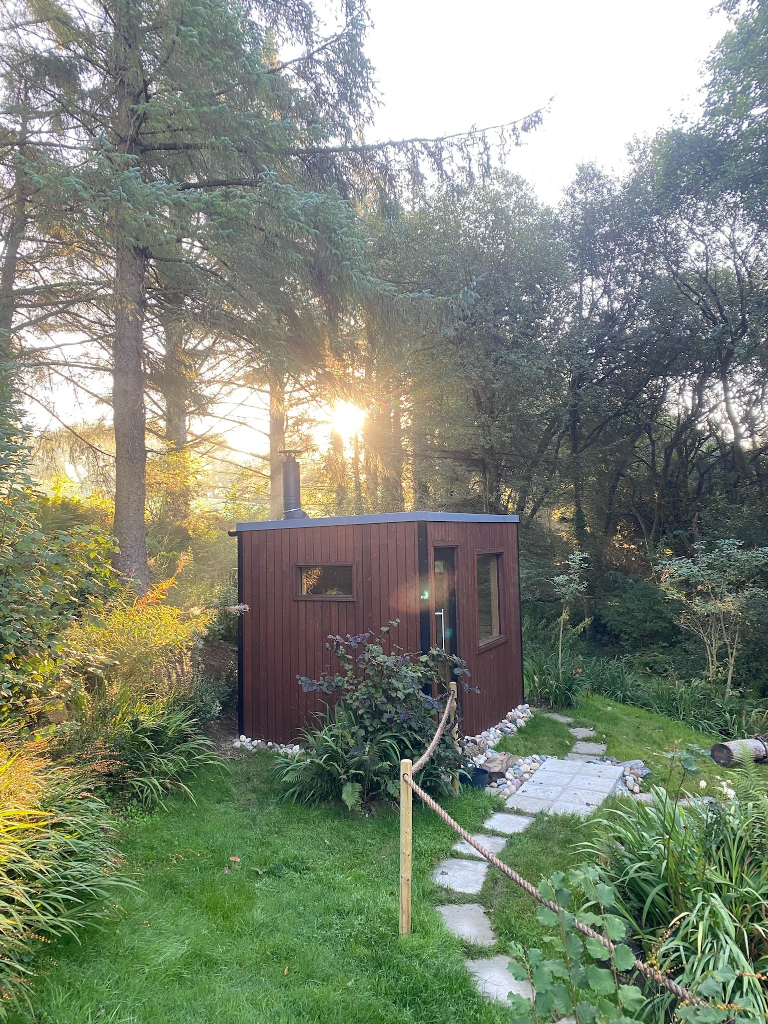 Small wooden cabin surrounded by greenery and trees with sunlight shining through the branches.