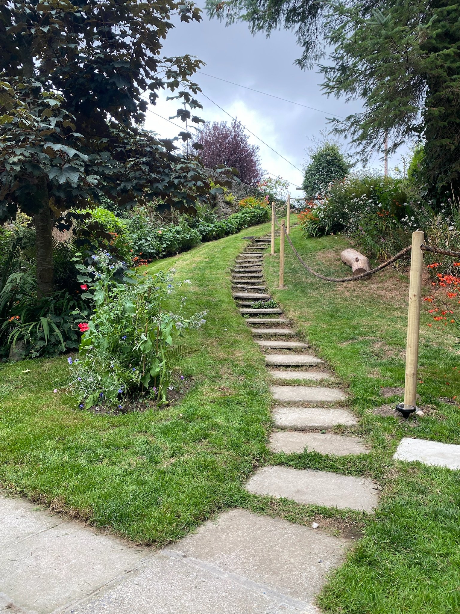 Stone pathway with steps leading uphill through a green garden with trees, flowers, and wooden posts connected by rope.