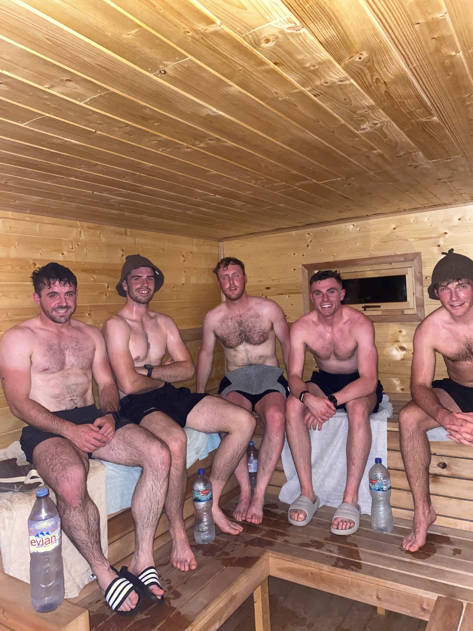 Five shirtless men sitting in a wooden sauna room with water bottles on the floor, two wearing hats and one wearing slides.
